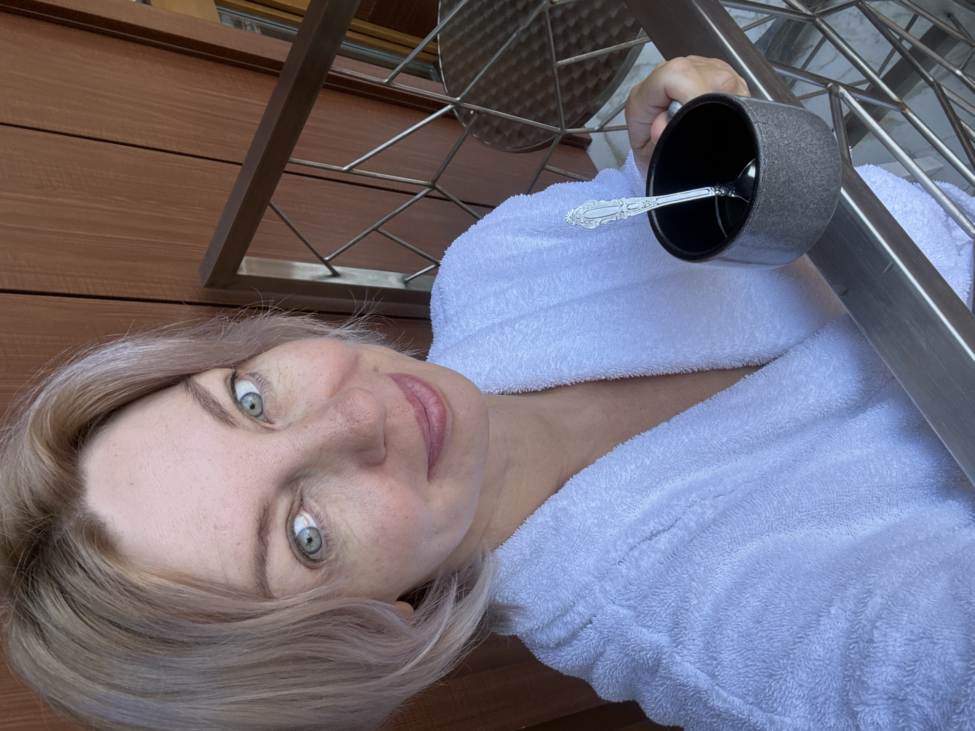 A woman, appearing to be in her 40s, with short, light blonde hair and green eyes, poses for a selfie on a balcony. She is wearing a white bathrobe and holding a dark mug with a spoon. The background consists of brown wooden paneling and a metal railing. The overall setting suggests a hotel room or apartment in Estosadok, Russia, possibly a resort area, given the balcony and the visible railing pattern. The lighting is natural and bright, indicating it is daytime. The woman's expression is relaxed and she is smiling faintly.
