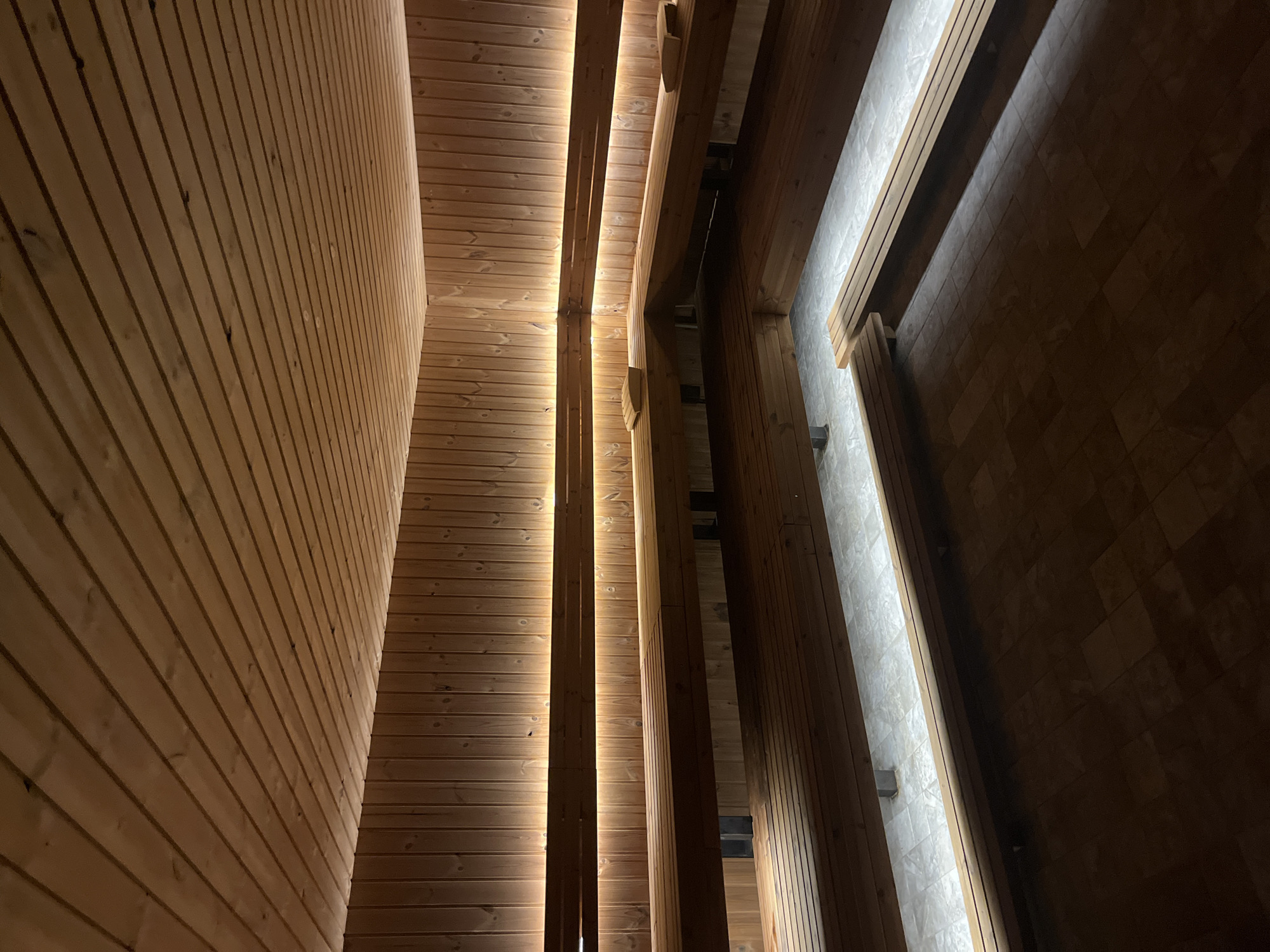 The image shows an interior view of a building, featuring wooden walls and structural elements. The walls are constructed with horizontally aligned wooden planks. Vertical wooden beams support the ceiling and possibly other levels above. There are light strips integrated between the beams, providing illumination to the space. The lighting is warm, creating an ambient atmosphere. The building might be located in Sofia, Bulgaria. The image focuses on the architectural details and lighting design. There are no people visible in the photograph.