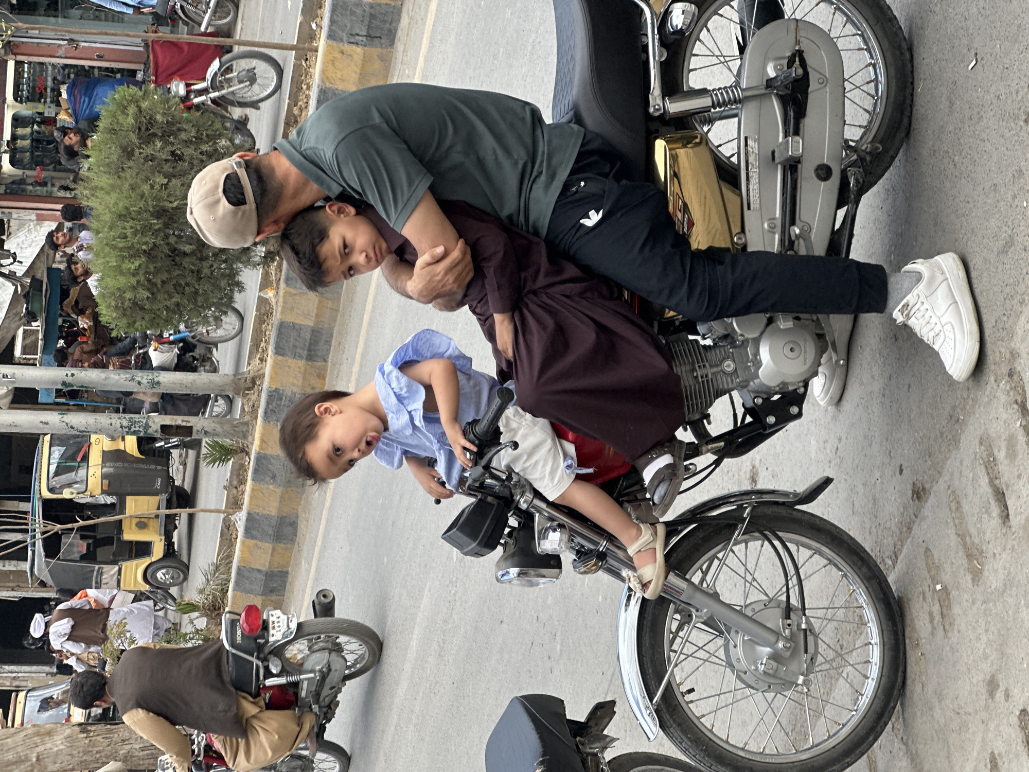 A medium shot shows a man and two children on a motorcycle in Quetta City Tehsil, Pakistan. The man, wearing a green shirt, dark pants, and a baseball cap, is seated on the motorcycle, holding a boy in a brown outfit close to him. In front of them, a younger child in a light blue shirt and white shorts sits on the motorcycle's handlebars. The motorcycle is silver with a black seat. The background includes a street with other motorcycles, pedestrians, and a yellow auto-rickshaw. There is a shopfront visible behind a tree. The road has a black and yellow striped curb.
