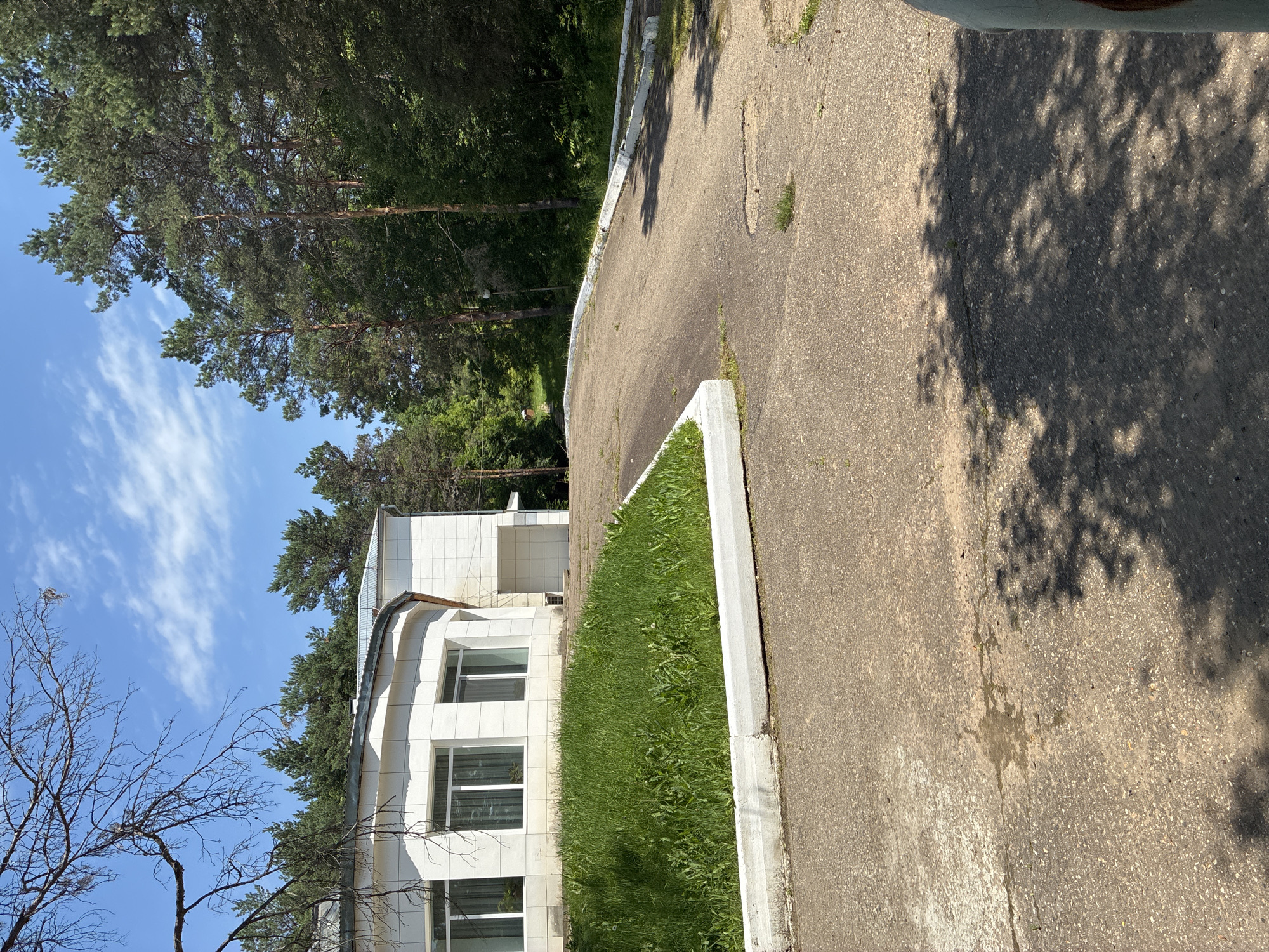 The image depicts an exterior view of a white tiled building with large windows, surrounded by lush green grass and tall pine trees under a clear blue sky with scattered white clouds. A paved asphalt pathway curves in front of the building, leading towards the trees and a gently sloping terrain. The scene appears to be a serene outdoor setting, possibly a park or a residential area, located in the city of Gornye Kljuchi, Russia. The lighting suggests it is daytime, with sunlight casting shadows on the pathway. There are no people or visible activities in the frame.