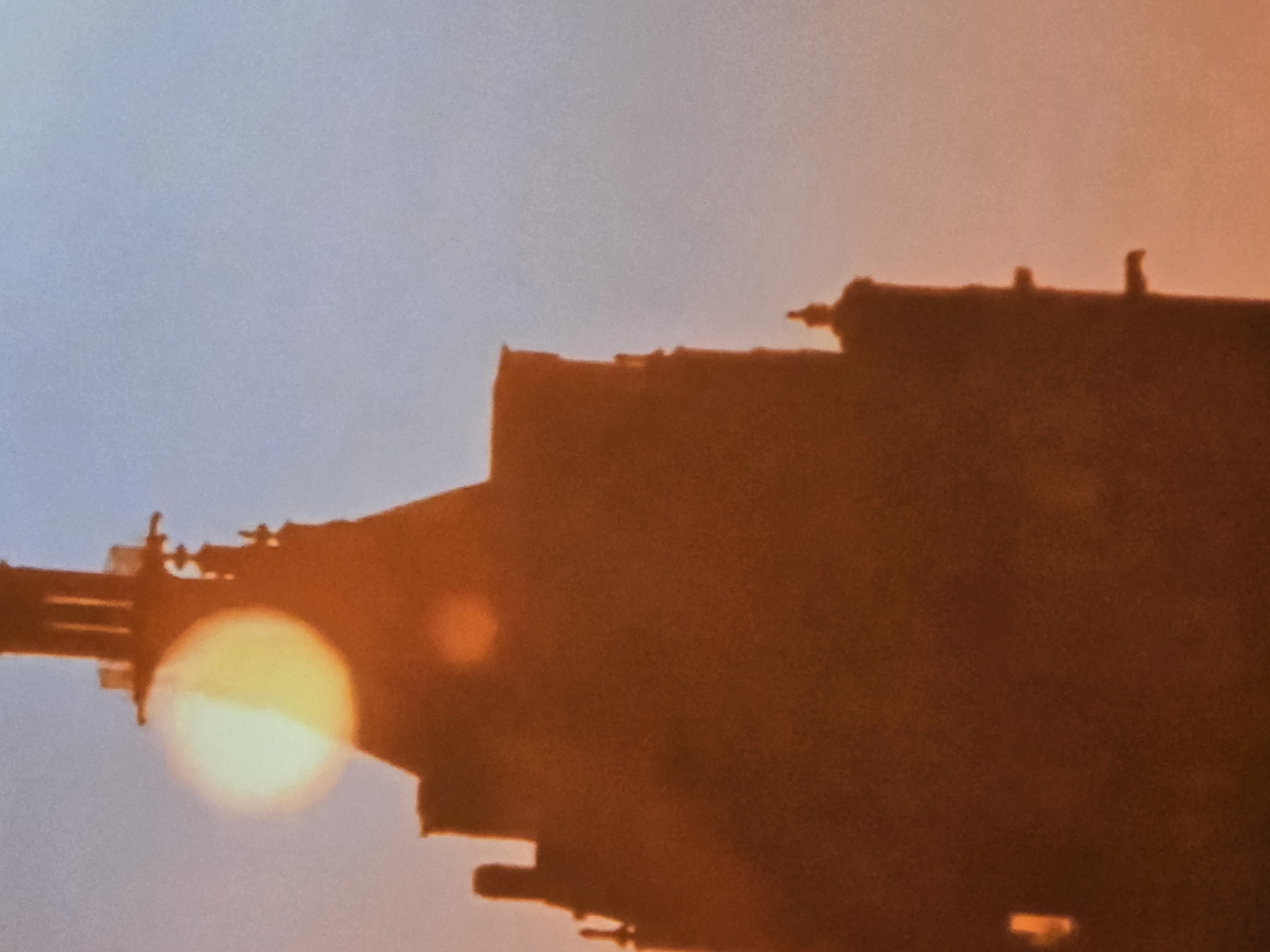 The image shows the silhouette of a building against a sunset or sunrise sky, with a bright light (possibly the sun) appearing to be obscured by part of the building's structure. The building's outline is complex, with various protrusions and levels. The details of the building are not clear due to the silhouette effect. The colors range from light blue in the upper left to orange and red near the horizon. Location is unknown.