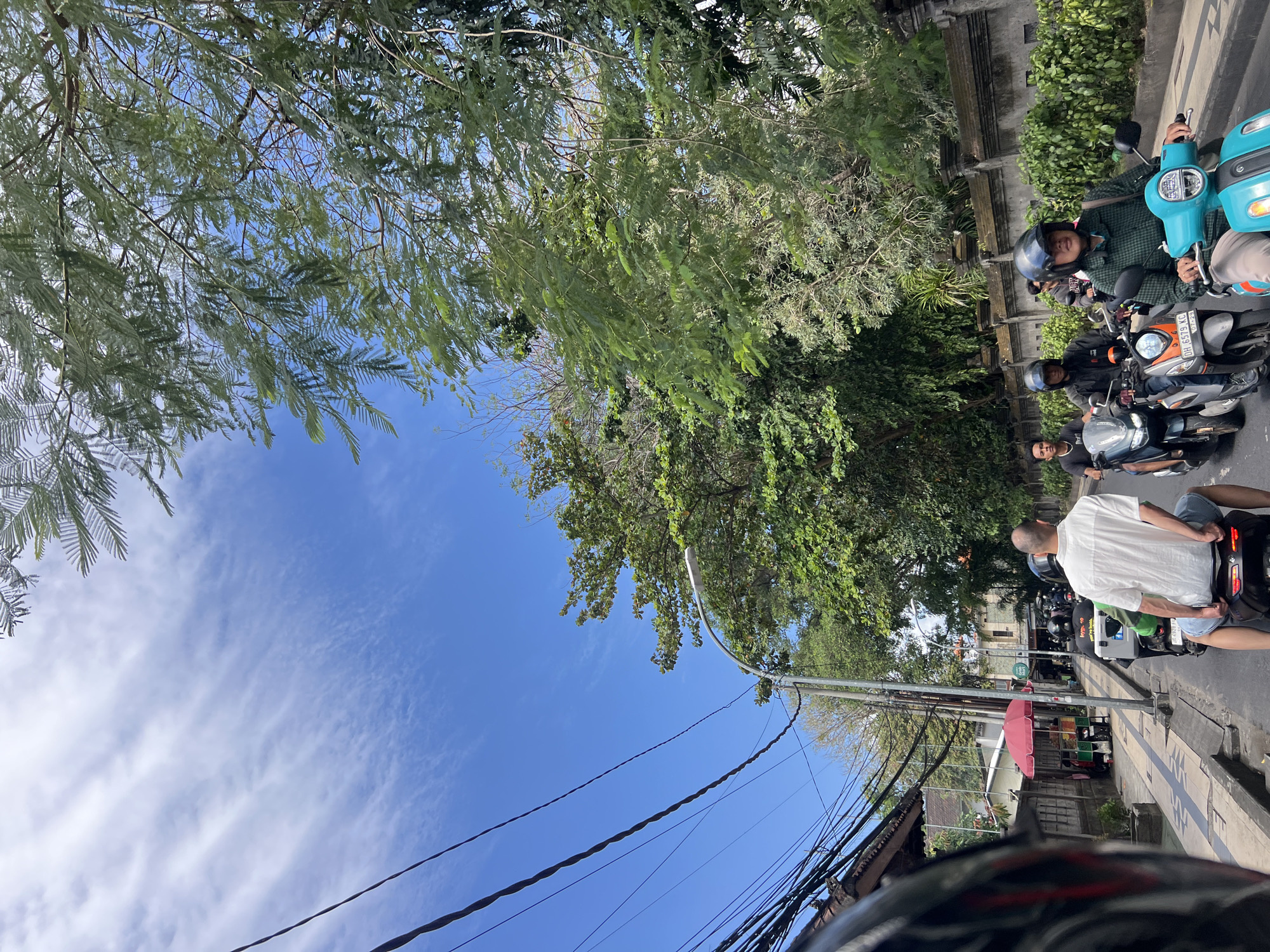 The image depicts a street scene in Kuta, Indonesia, on a bright, sunny day with a blue sky dotted with wispy clouds. A line of motorcycles navigates the road, with several riders visible. In the foreground, a person wearing a white shirt and blue shorts is seated on a motorcycle, seen from behind. Other riders are also on motorcycles, some wearing helmets. The street is lined with buildings and lush green trees. Power lines are strung overhead. To the left of the road, a small food stall with a red umbrella is visible. The overall atmosphere suggests a busy, common day in the bustling city.