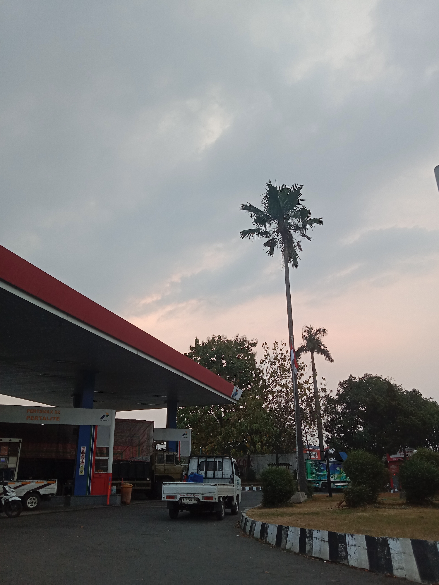 This image depicts a gas station under an overcast sky, likely at dusk. The gas station features a red-trimmed canopy over multiple fuel pumps, with signs indicating "Pertamax 92" and "Pertamax Lite." A white motorcycle and a white pickup truck are parked at the station. In the background, several larger trucks are visible. Two prominent palm trees stand tall against the sky, one significantly taller than the other, with a small Indonesian flag attached to the taller tree's trunk. Trees and green bushes surround the area, and a curved concrete barrier with black and white stripes lines the edge of the paved area. The scene suggests a roadside location within a city, possibly Semarang, Indonesia, given the typical appearance of gas stations and vegetation in that region. There are no people visible in the image.