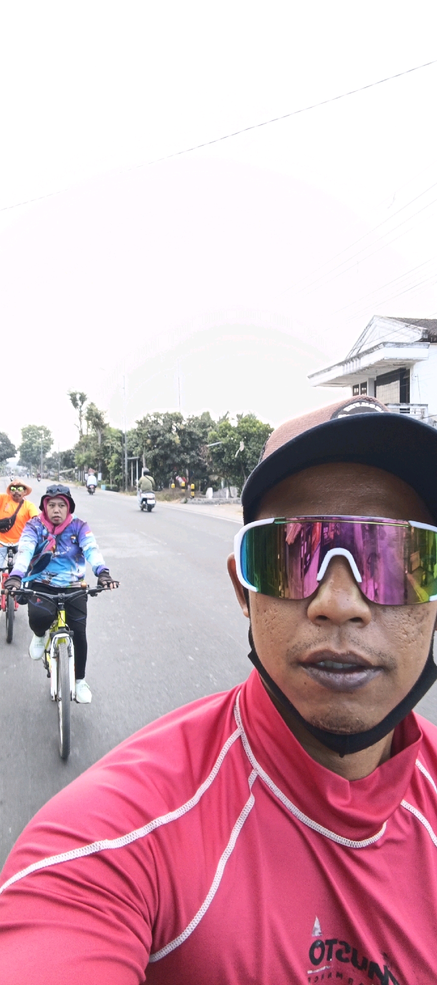 The image shows a group of people cycling on a road in Kasiyan, Indonesia. In the foreground, a person is taking a selfie, wearing a red long-sleeved shirt, a black cap, and large, multicolored cycling glasses. Behind them, a woman in a pink headscarf and a colorful long-sleeved shirt is cycling on a yellow bicycle. Further behind, another person in an orange shirt and a hat is also cycling. Several motorbikes are visible in the background, along with trees and a building. The road appears to be paved, and the sky is overcast.