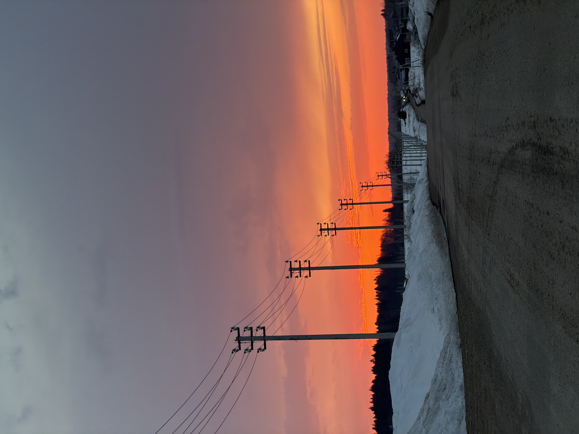 The image shows a roadside scene at sunset in a snowy environment. On the right is an asphalt road. Parallel to the road on the left is a layer of packed snow. Further left are telephone poles, with wires stretching diagonally across the image. Beyond the telephone poles is a forest line and a vibrant sunset sky of orange and purple hues. The location could be near Izhevsk, Russia, due to the landscape's characteristics and prevalence of snow. There are no people visible.