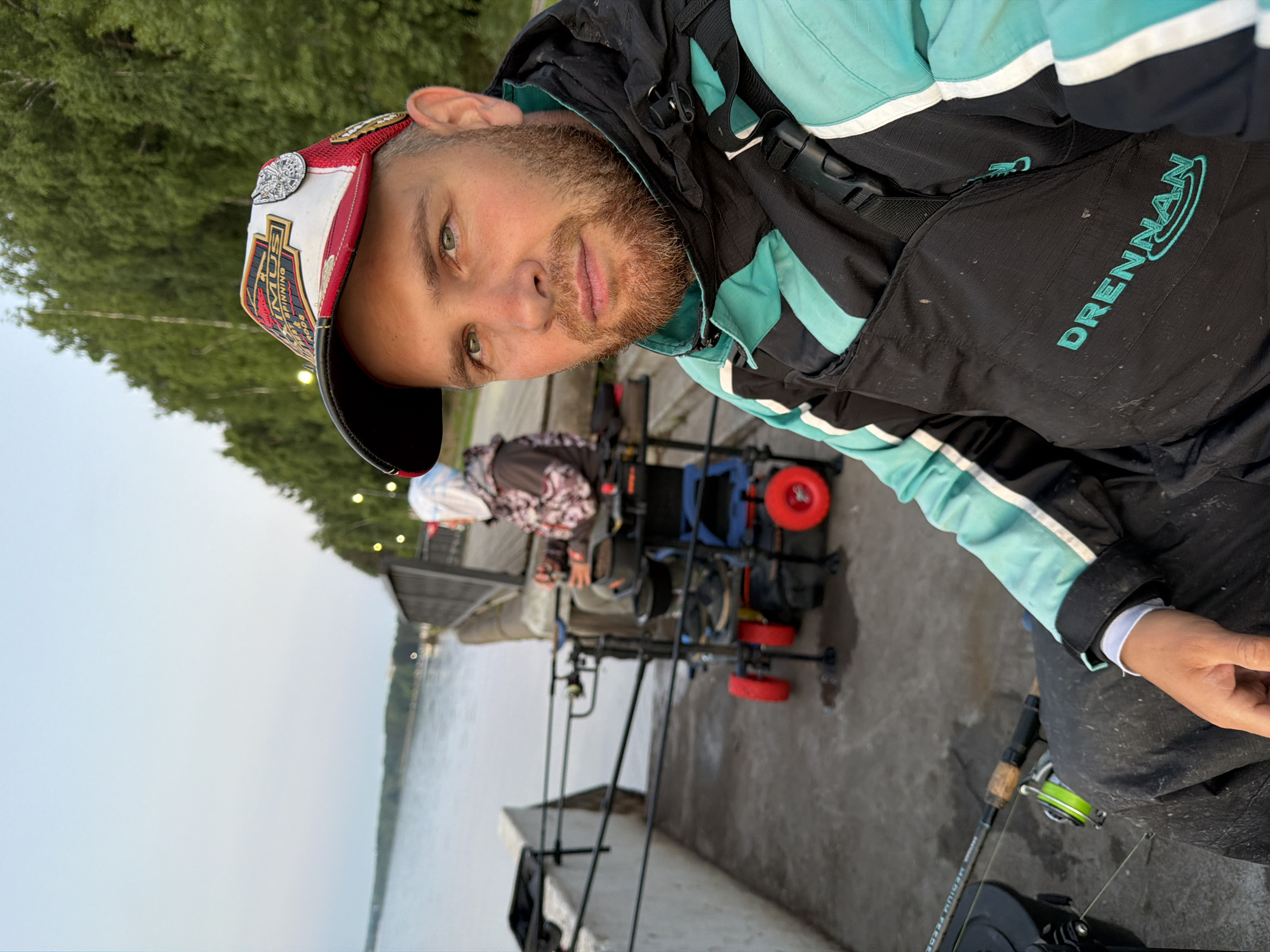 A man with a beard and a baseball cap is sitting on a concrete pier, fishing. He is wearing a black and turquoise fishing jacket and bib overalls with the brand "DRENNAN" visible. His fishing rod and reel are in front of him. In the background, another person is seated in a fishing chair, also fishing, with fishing rods extending towards the water. The scene appears to be at dusk, with a calm body of water next to the pier and trees lining the opposite bank, likely in the city of Chaikovsky, Russia, possibly along the Kama River.
