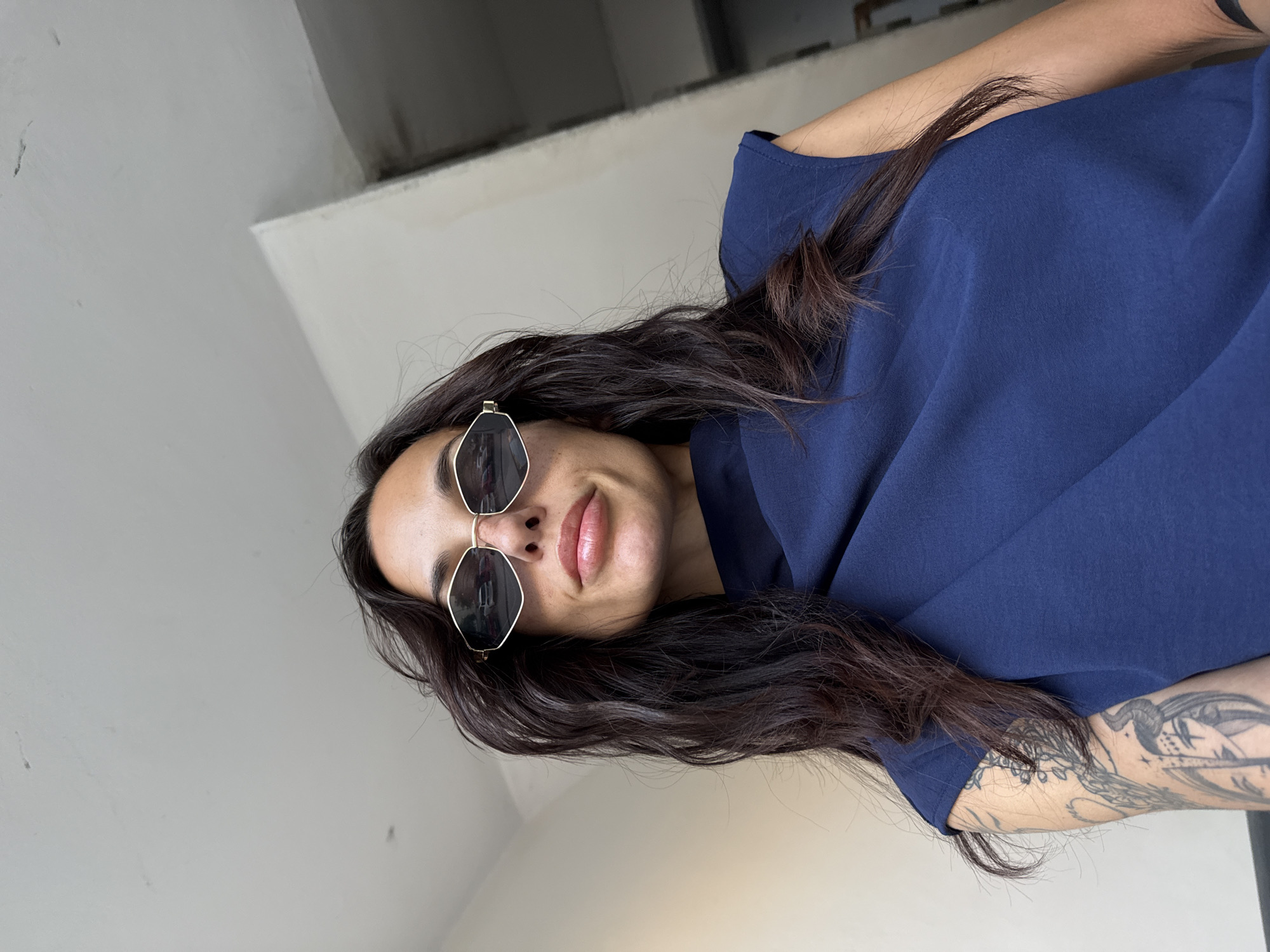 A medium shot depicts a woman with long, dark hair wearing sunglasses and a blue t-shirt. She is indoors in what appears to be a hallway or building interior with neutral-colored walls. The woman also has a tattoo on her arm. It's not possible to tell from the image if the location is indeed in Pleven, Bulgaria.