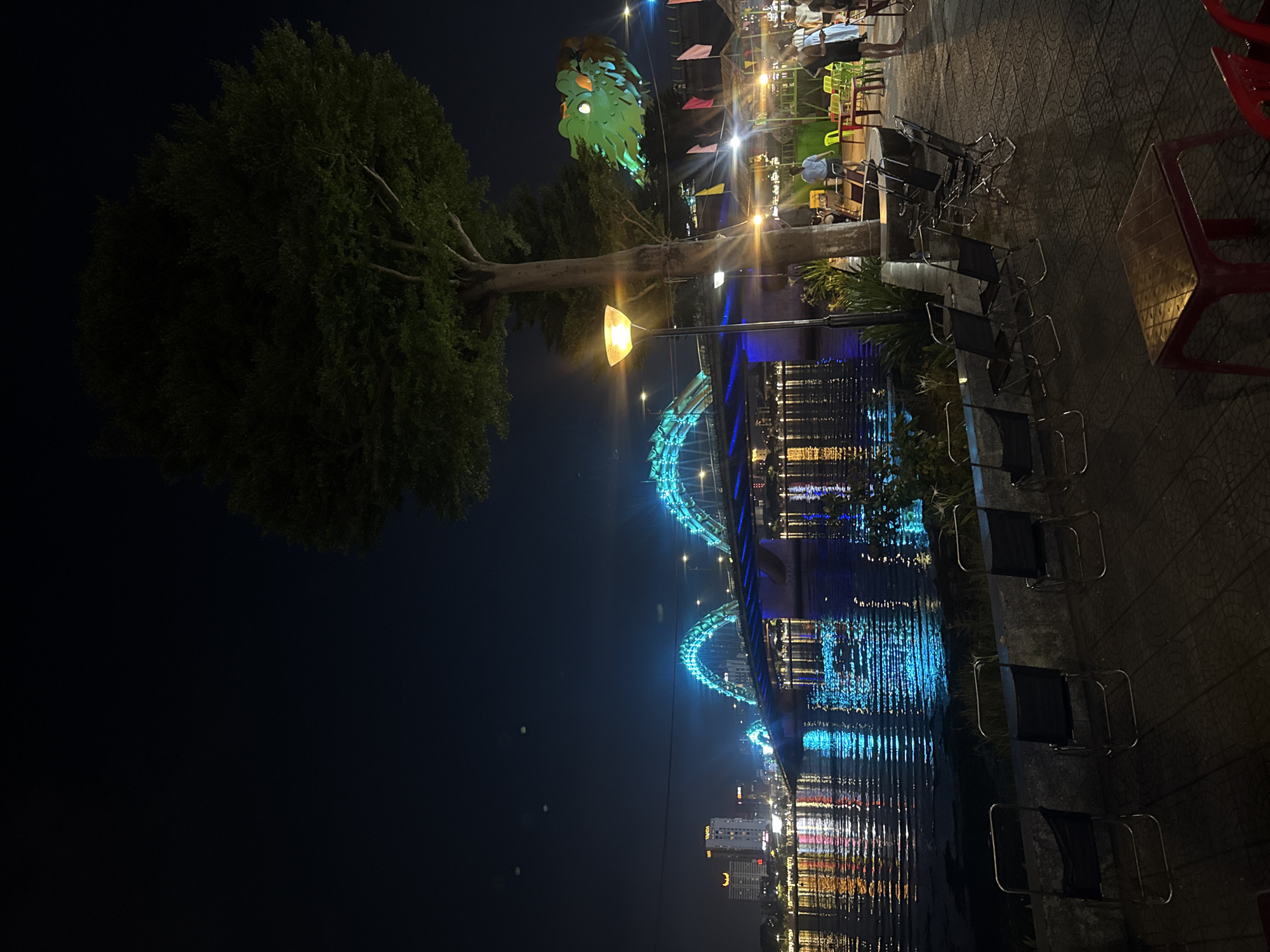 Captured at night, the image depicts a city scene in Unknown, Unknown, focusing on a waterway reflecting city lights. A prominently lit bridge arches across the water, its blue lights mirroring in the dark water below, where the reflections of city buildings are visible. The foreground features a tree on the left and an outdoor dining area on the right. The dining area is furnished with chairs and tables, suggesting a restaurant or café atmosphere, and some people are seated and walking, though details are not clear. Above the dining area, a unique decorative structure, possibly a sculpture or illuminated art installation, adds to the ambience. The ground surface is paved. The lighting is artificial, with streetlights and the bridge lights contributing to the overall glow.