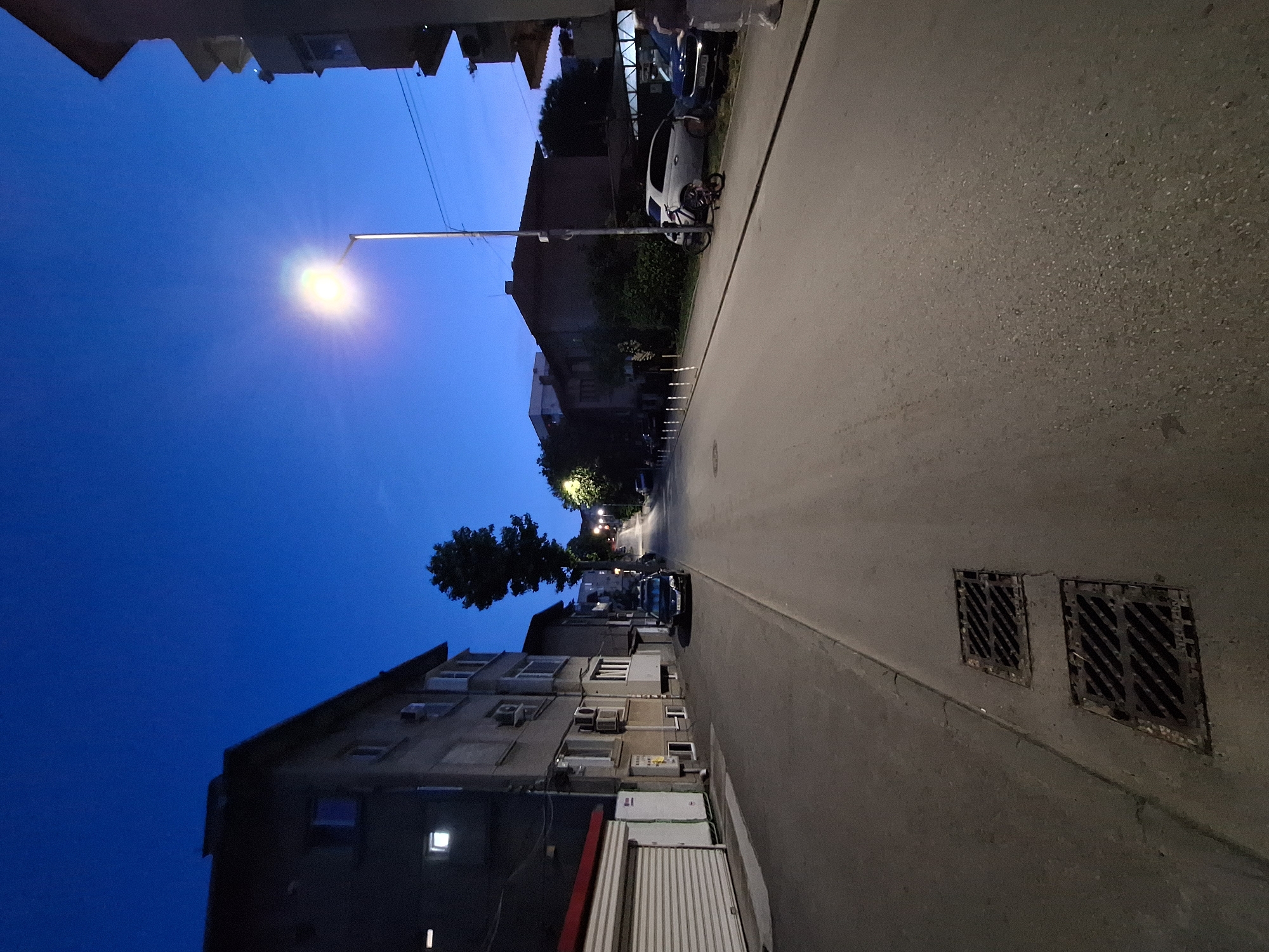 The image depicts a street scene in Varna, Bulgaria, at dusk. The sky is a deep blue, indicating the transition from daylight to night. A street lamp illuminates the asphalt road, which slopes gently upwards. On the left side of the road, a multi-story building with several windows and air conditioning units is visible, along with a closed garage door. Further down the street, more buildings and trees can be seen. On the right side of the road, parked cars and a bicycle are present. Two manhole covers are situated on the sidewalk in the foreground. No people are clearly visible in the image. The environment appears to be a residential or mixed-use urban area.