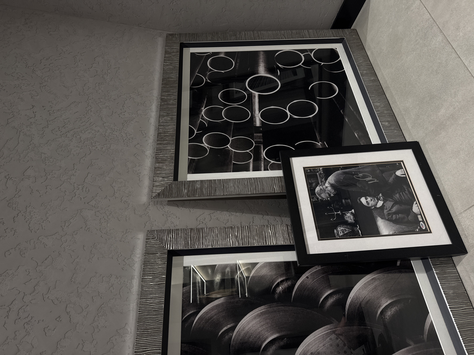 The image displays an indoor corner featuring three framed photographs, predominantly in black and white, against contrasting wall textures. The scene presents a monochromatic aesthetic with various shades of grey, black, and white.
On the left, the wall has a rough, stucco-like texture in a light grey tone. On the right, a smooth, lighter grey wall or tiled surface forms the adjacent side of the corner.
Two large, vertically stacked pictures are mounted on the wall. The top frame encloses an abstract black and white photograph depicting multiple circular objects, possibly pipes or tubes, arranged against a dark background. The frame itself is distinctive, with a metallic, striped texture in grey and silver tones.
Below it, a second large picture, framed identically, is partially visible. This black and white image shows a collection of stacked, rounded objects, suggestive of industrial components like large gears or turbine blades. Reflections are visible on its glass surface from indoor lighting.
Leaning against the upper large frame and partially obscuring the lower one is a smaller, rectangular framed photograph. This black and white portrait features two men. An older man, mostly seen from the back or side, looks down towards a younger man seated in an armchair. The younger man, dressed in a suit and tie, looks directly at the viewer. The older man appears to wear a light-colored sweater with faint text that resembles "G C" or "O C." A small white sticker with the text "PSA" and a number (e.g., "8") is visible on the white matting of this picture, likely indicating authentication or grading. This smaller frame is a simple black design with a white mat.
The overall impression is of a contemporary and well-curated interior space, possibly a hallway or lounge area, with a focus on sophisticated, monochrome art. No specific visual cues within the image directly indicate its location in Dubai, United Arab Emirates, beyond a generally upscale interior design.