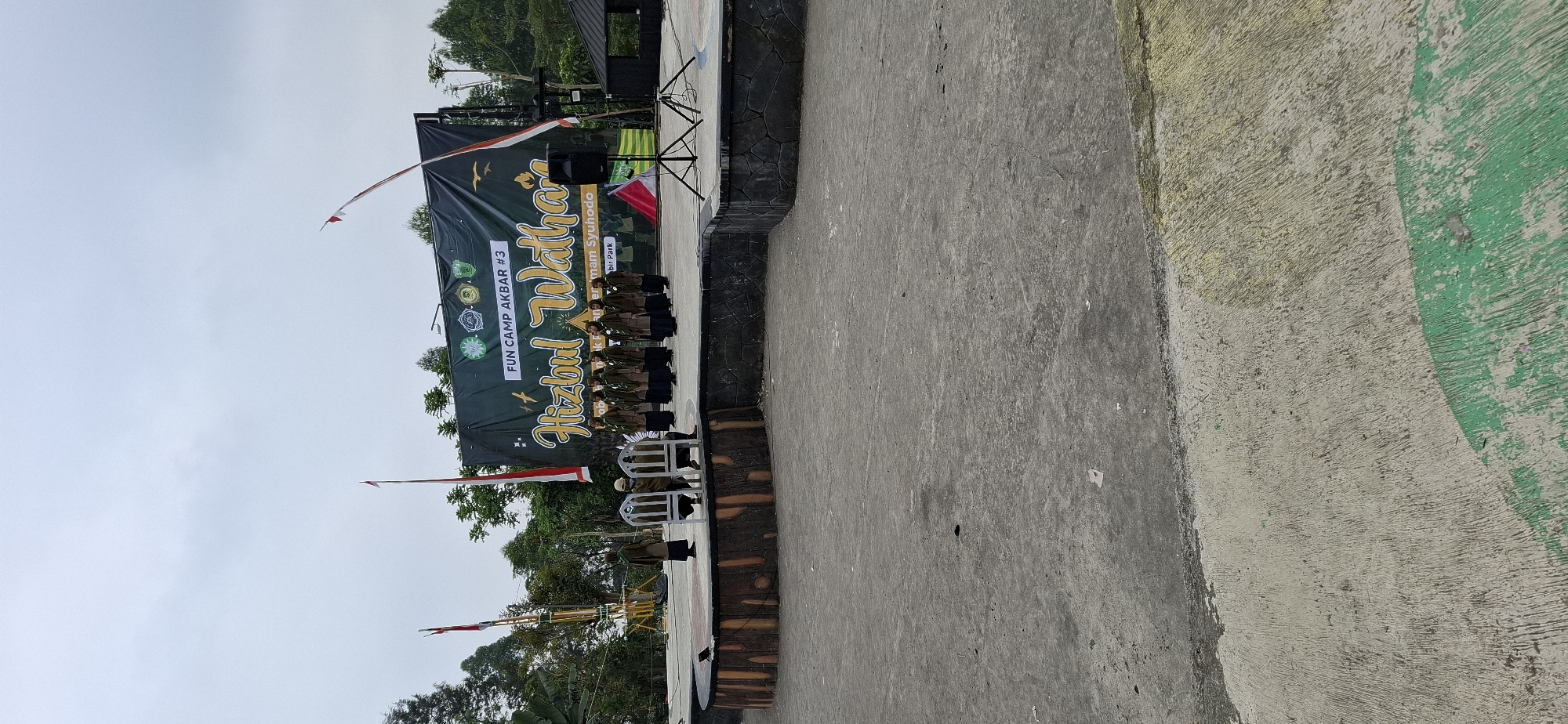 An outdoor stage area is set up for an event. A large, dark green banner with yellow text and logos hangs behind the stage. The text on the banner appears to read "FUN CAMP AKBAR #3" and "Hizbul Wathan." A group of approximately ten people, dressed in matching brown uniforms and headscarves, stand on the stage, facing towards the left of the frame. Two chairs are placed on the stage, unoccupied. The stage itself is a semi-circular wooden platform with a stone border. To the left of the stage, two tall, yellow poles with flags are visible. In front of the stage, a concrete ground area extends outwards. A black speaker on a tripod is positioned on the right side of the stage. The sky is overcast. The image appears to be taken in the city of Karang, Indonesia.