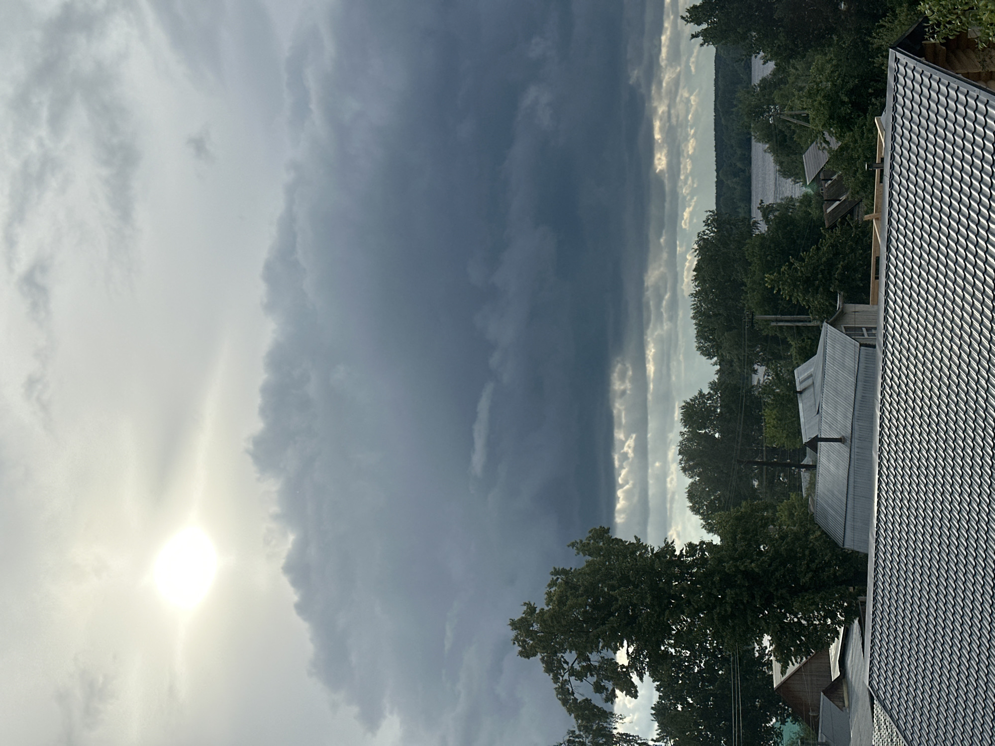 The image depicts a dramatic landscape featuring a village or settlement in the foreground, bordering a large body of water, all under a dynamic sky. The sky dominates the upper portion, showcasing a stark contrast between a brilliant, almost blinding sunburst on the left and extensive, dark, heavy storm clouds spanning the center and right. Lighter, wispy clouds are visible around the sun.
In the midground, a wide river or lake extends horizontally, its surface reflecting the overcast sky and appearing somewhat dark, with occasional glints of light. The banks of the water body are densely forested with lush green trees. Several houses or buildings with sloped roofs, some appearing to be corrugated metal, are nestled among the trees and along the water's edge. The closest structure in the bottom right features a distinctive, patterned metal roof. Power lines and poles are also visible, indicating infrastructure.
No human activity is present. The scene strongly suggests a period of dramatic weather, possibly an impending or clearing storm, with the sun breaking through intense cloud cover. This could be during late afternoon or early evening. The overall atmosphere conveys powerful natural beauty and changing meteorological conditions. There is no visible text in the image. The setting is consistent with a rural or semi-rural area near a large body of water, fitting the location context of Chaikovsky, Russia, and the Kama River.