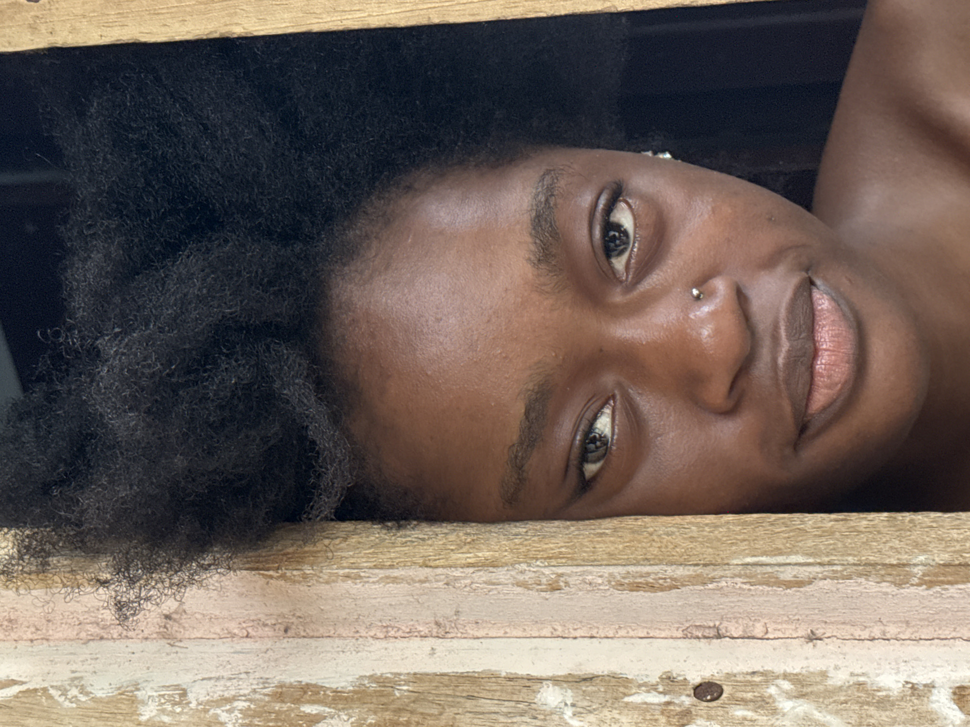 A close-up, head-and-shoulders shot features a young Black woman with dark, voluminous natural hair. She is positioned to the right of a weathered wooden frame, with one side of the frame obscuring the left edge of the image. Her face is turned slightly towards the viewer, with her eyes looking directly ahead. She has a small nose stud, and a faint smile plays on her lips. The background is dark and indistinct, suggesting an indoor setting or a shaded outdoor area. The wooden frame shows signs of age and wear, with peeling paint and visible grain. The lighting is soft, highlighting the contours of her face and casting subtle shadows. The image appears to have been taken in Ado, Nigeria.