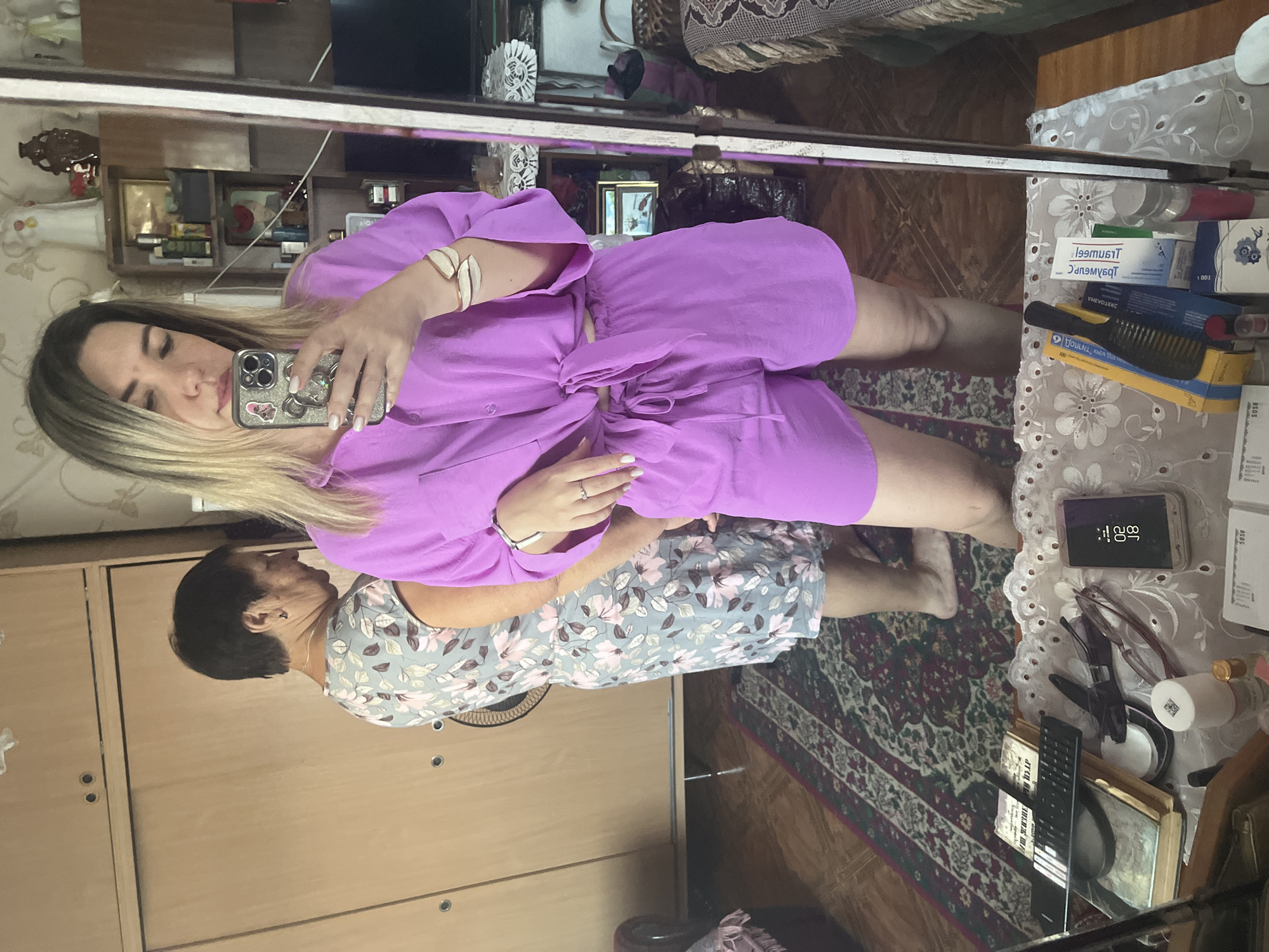 A young woman is taking a selfie in a mirror, wearing a pink tie-front top and matching shorts. To her left, an older woman in a floral dress stands with her back to the camera. The scene appears to be an indoor setting, possibly a bedroom or living room, with wooden furniture, a patterned rug on the floor, and various objects visible on shelves and surfaces in the background.