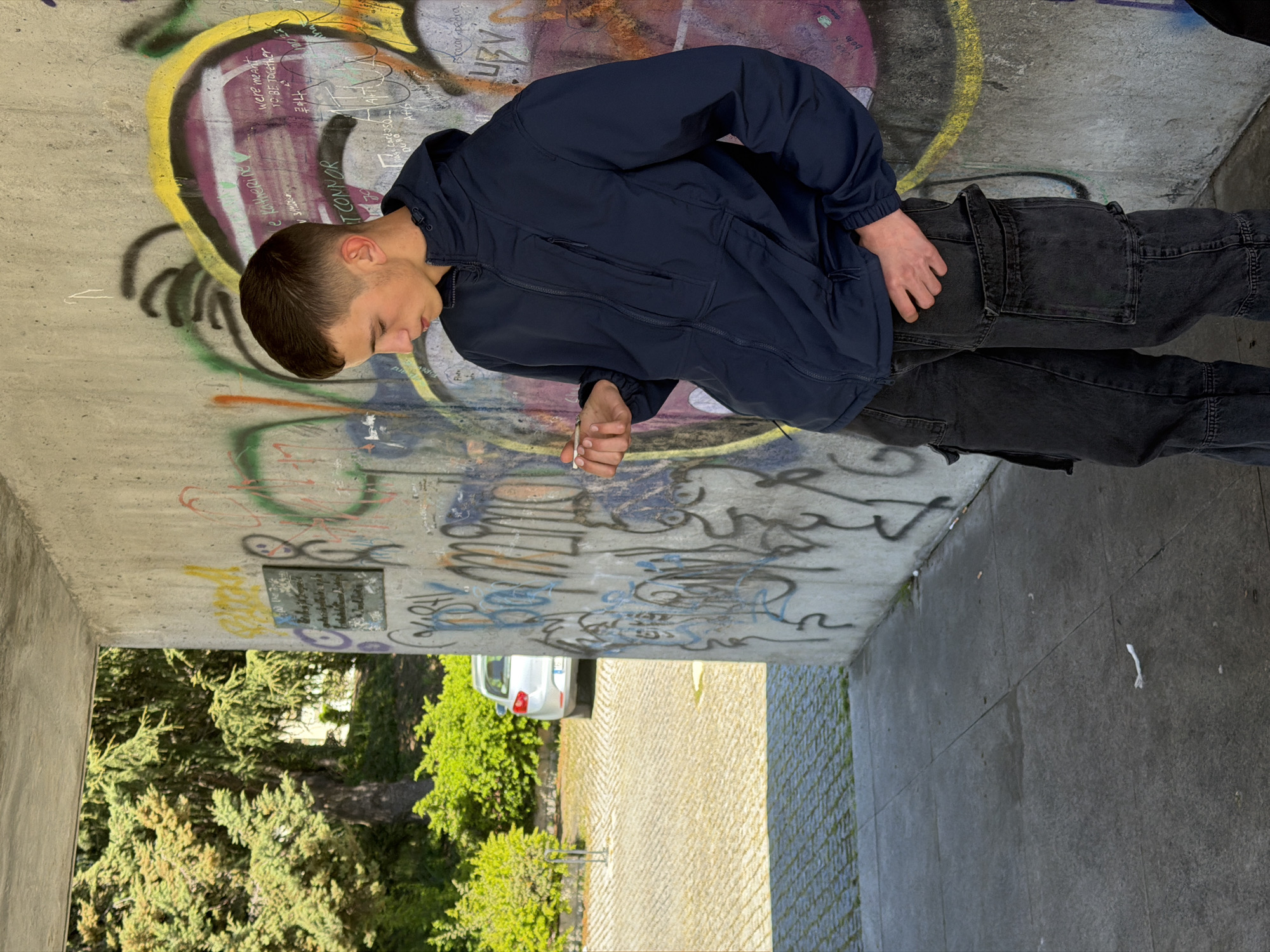 The image captures a young adult male standing under a concrete structure, likely an underpass or sheltered walkway, in Vila Real, Portugal, during daylight hours. The male, with short dark hair, wears a dark blue hooded zip-up jacket and dark grey cargo pants. He leans against a textured concrete wall heavily covered with multi-colored graffiti, displaying various spray-painted texts and abstract designs. A small, dark rectangular plaque is affixed to the wall to his left. He is looking down at a small, white, cylindrical object, consistent with a hand-rolled cigarette or joint, held between the thumb and forefinger of his left hand; his right hand is in his pants pocket. The immediate ground consists of grey concrete paving slabs with minimal debris. Beyond the concrete structure, a paved area with tan cobblestones is visible, where a silver/white hatchback car is partially parked, surrounded by dense green trees and bushes.