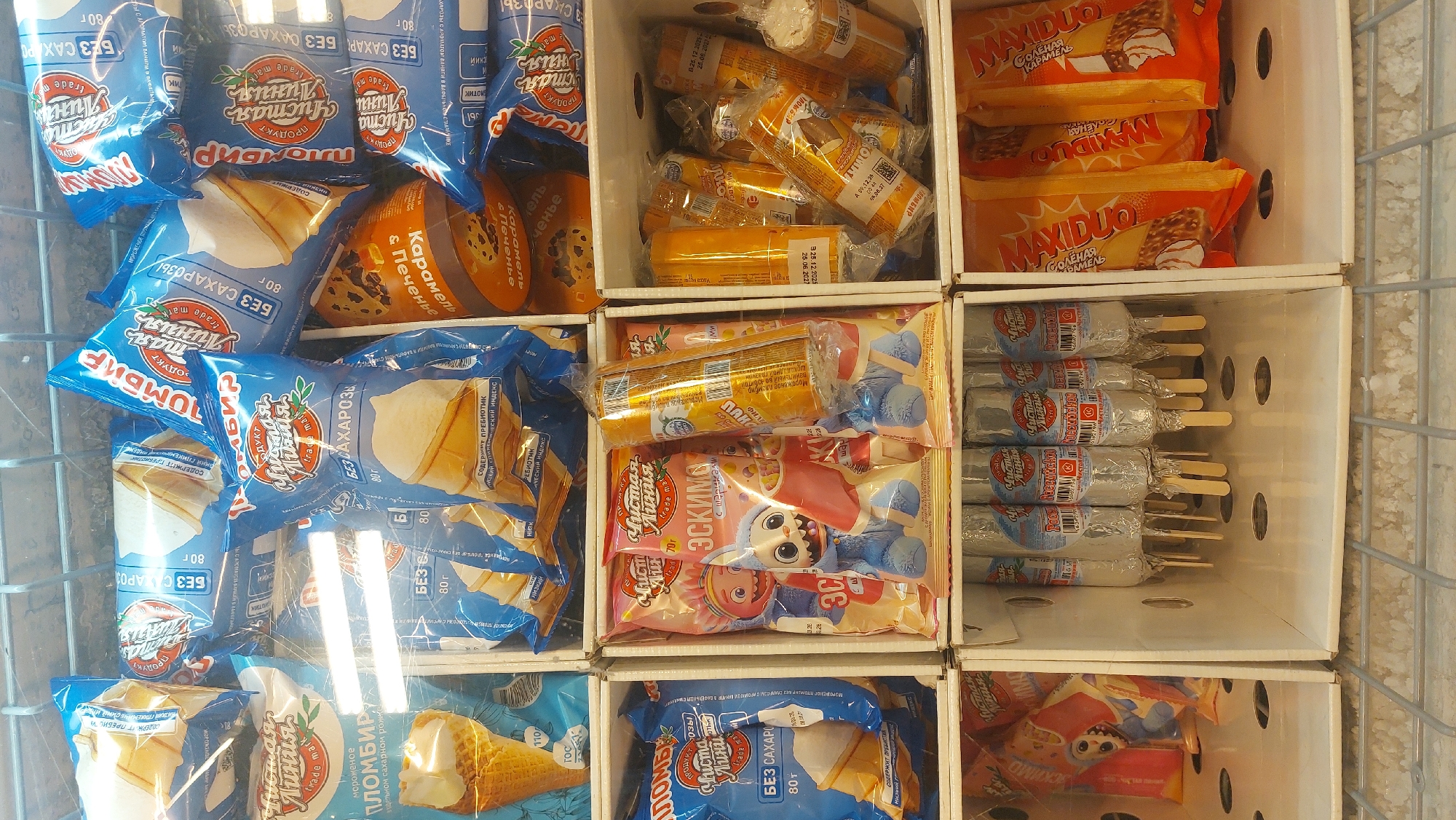The image shows a freezer display case containing various ice cream products. The brands visible include "Chistaya Liniya" (Чистая Линия) and "Maxiduo." The ice cream comes in several flavors and forms, such as "Plombir," caramel with cookies, and ice cream on a stick. The packaging indicates "Bez Sakhara" (Без сахара), meaning "without sugar." The packaging is in Russian, indicating that the location is likely in Russia. Given that the OCR text extracted also indicates the brands are Russian, it seems the product is being sold in Russia, possibly Novosibirsk.