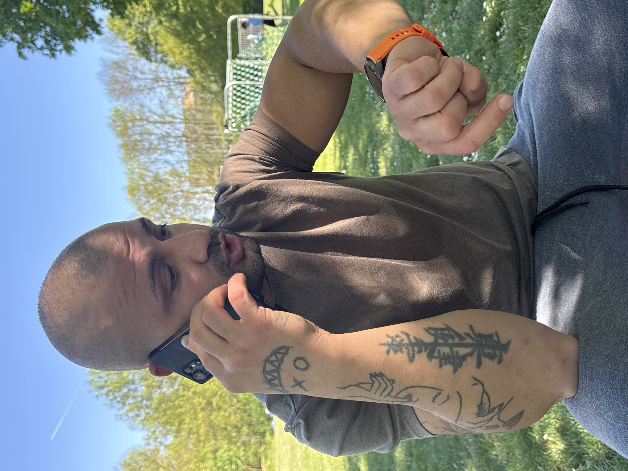 A bald or short-haired man is captured outdoors, talking on a black smartphone held to his left ear. He is wearing a dark brown t-shirt and grey sweatpants. His right arm is bent, revealing an orange-strapped watch on his wrist, and his hand is partially clenched.
The man has several tattoos on his left arm: on his forearm, there is a design featuring "XO" alongside a stylized grinning mouth with jagged teeth. Further up his left arm, there are other abstract designs, including one that appears to depict stylized trees or a symbolic script.
The scene is set in a sunny, bright outdoor location, likely a park or a grassy field in Ditzingen, Germany. The background features lush green grass, deciduous trees, and a clear blue sky with a visible contrail. A metal structure, possibly part of a goal post or play equipment, is visible amidst the trees. The strong sunlight suggests it is daytime. The man appears engaged in his phone conversation.