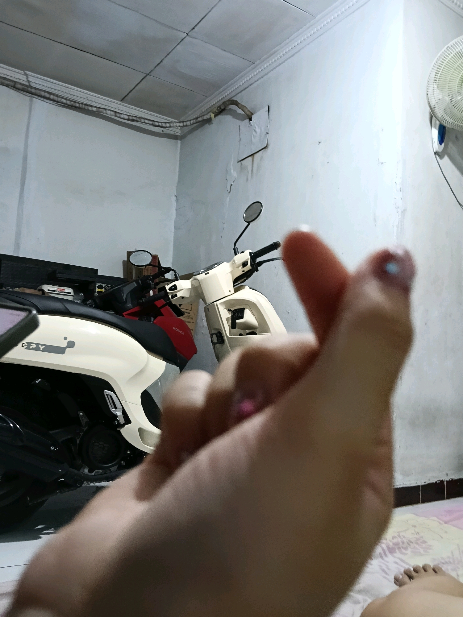 A close-up, blurry, indoor, eye-level medium shot shows a person's hands in the foreground, forming a Korean heart gesture. Behind the hands, a beige and black scooter is parked. The background features a white, textured wall with some peeling paint, a ceiling with visible tiles and wiring, and a white electric fan mounted high on the wall. The floor appears to be tiled, and a patterned bed is visible in the lower right corner. The image is shot in low light conditions, with the primary light source appearing to be from the right. The exact location within Jatimulya, Indonesia, cannot be determined from the visual information alone.