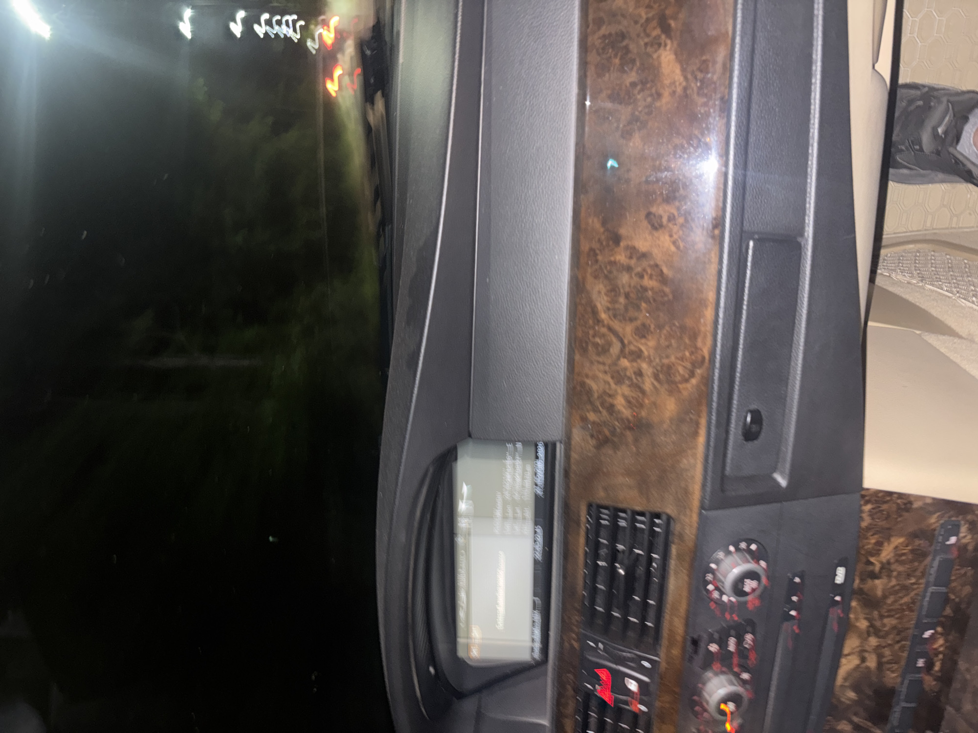 The image shows the interior of a vehicle, specifically the dashboard area. To the left, a screen displays information. Adjacent to the screen is a dark gray control panel, with what appears to be air vents, dials, and buttons. To the right of the control panel is a section with a wood grain finish. The exterior view through the windshield shows a dark environment with blurred lights, which appears to be an urban environment. The presence of greenery and blurred street lights suggests that the vehicle might be parked on a street within Plovdiv, Bulgaria. Due to the limited visual information, I cannot discern any more details about this scene.