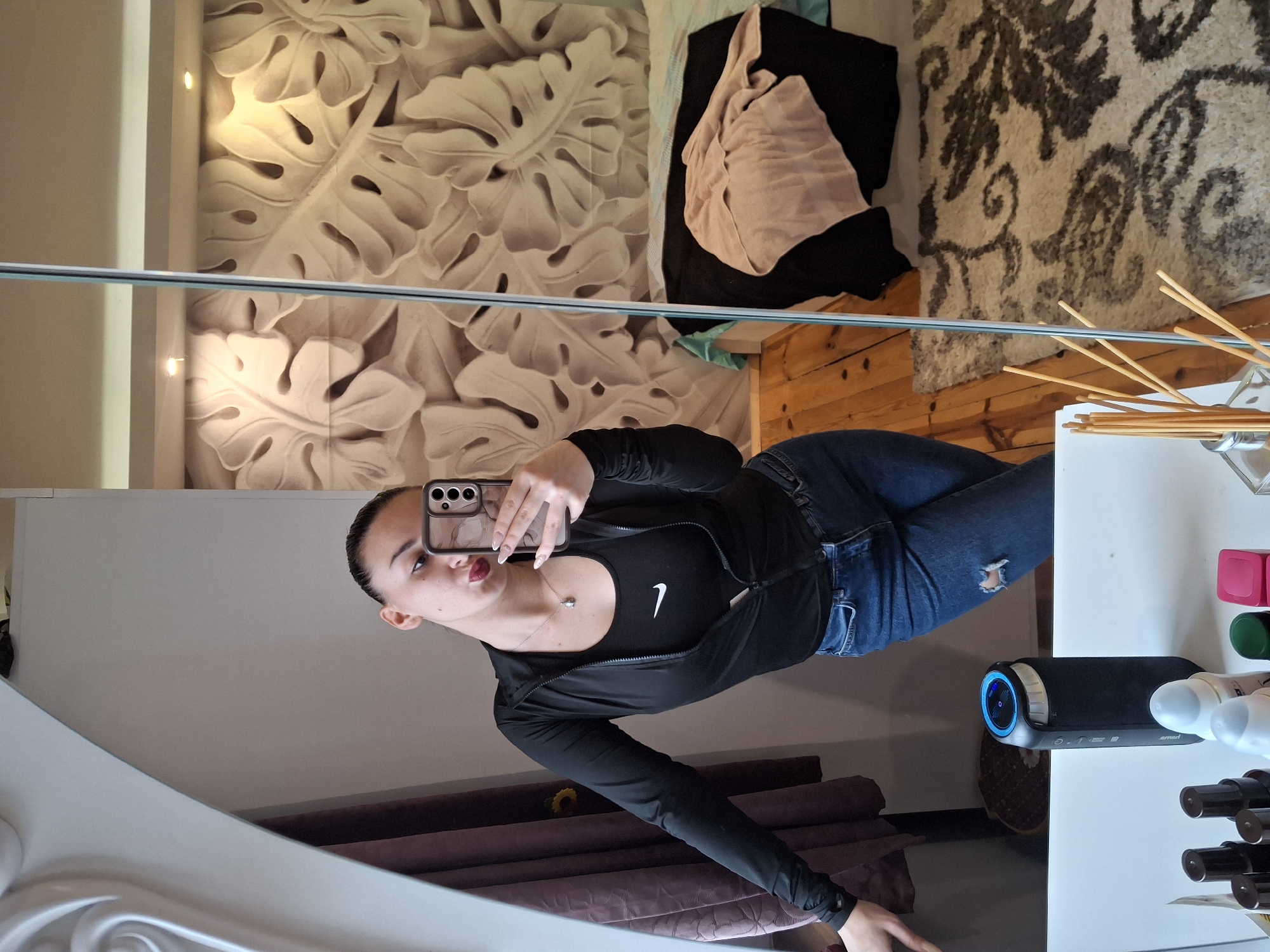 A young woman is taking a selfie in front of a mirror in what appears to be a bedroom. She is wearing a black Nike tank top, a black zip-up jacket, and blue jeans. The background features a wall with a raised leaf pattern, a bed with blankets, a rug, and a wooden floor. A table with various items, including a diffuser and a black speaker, is visible to the right. The scene appears to be indoors, possibly in a home located in an unknown area. Buhovo, Bulgaria is not mentioned in any of the information in the image.