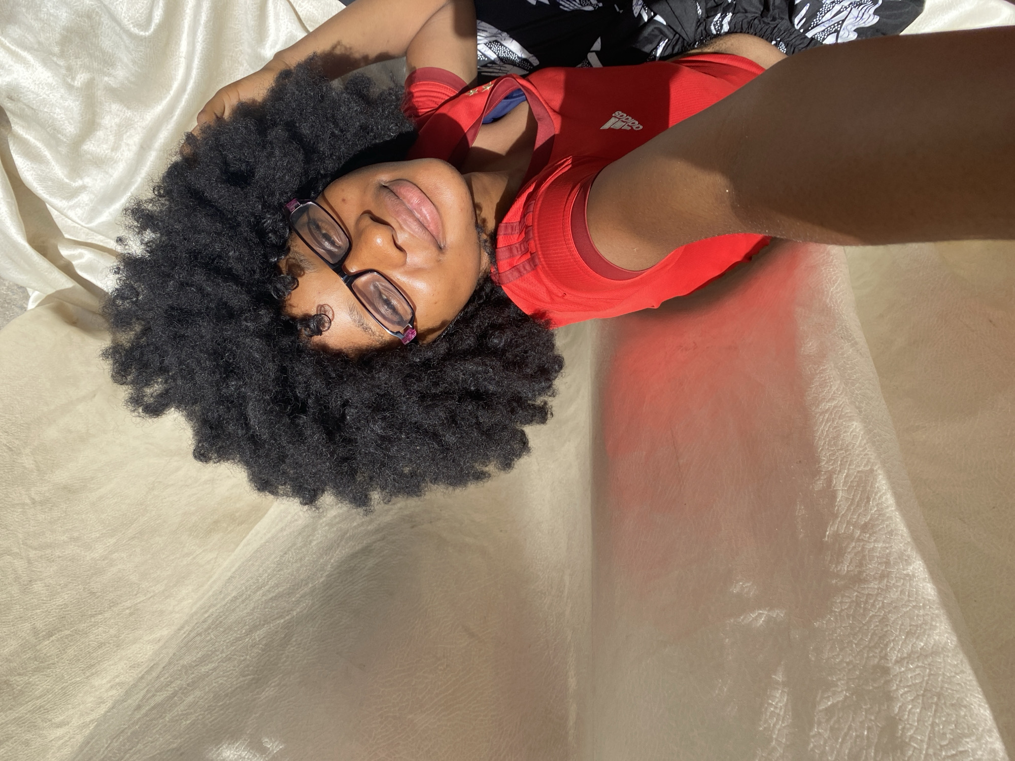 A dark-skinned person with black curly hair and glasses is lying on a light-colored sheet, wearing a red Adidas shirt and black and white patterned pants. The person is positioned at an angle to the camera, with their face and torso visible. The arm holding the camera is visible on the right side of the image. The background consists of the light-colored sheet with some shadows.
The location of Iranla, Nigeria is not discernible in the image.