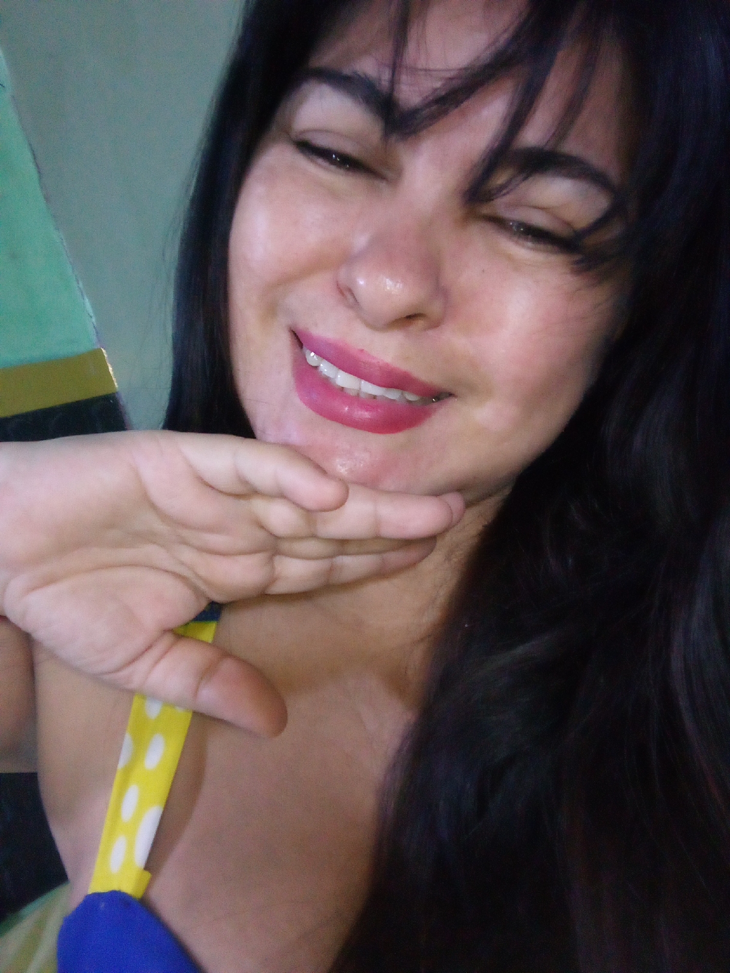 A close-up selfie shows a woman with dark hair, smiling broadly with her eyes squinted or closed. Her right hand is placed gently against her chin and lower jaw. She is wearing bright pink lipstick, which highlights her white teeth. Her hair is dark and falls around her face and shoulders.
Visible clothing includes a yellow strap with white polka dots, likely part of a top or swimsuit, which appears to be layered over a blue garment. The strap is visible near her shoulder and hand.
In the background, to the left, there's a soft green object or surface with a horizontal gold/yellow stripe. The overall setting appears to be indoors and casually lit. The woman's expression conveys happiness, amusement, or a playful mood. No text is visible in the image.