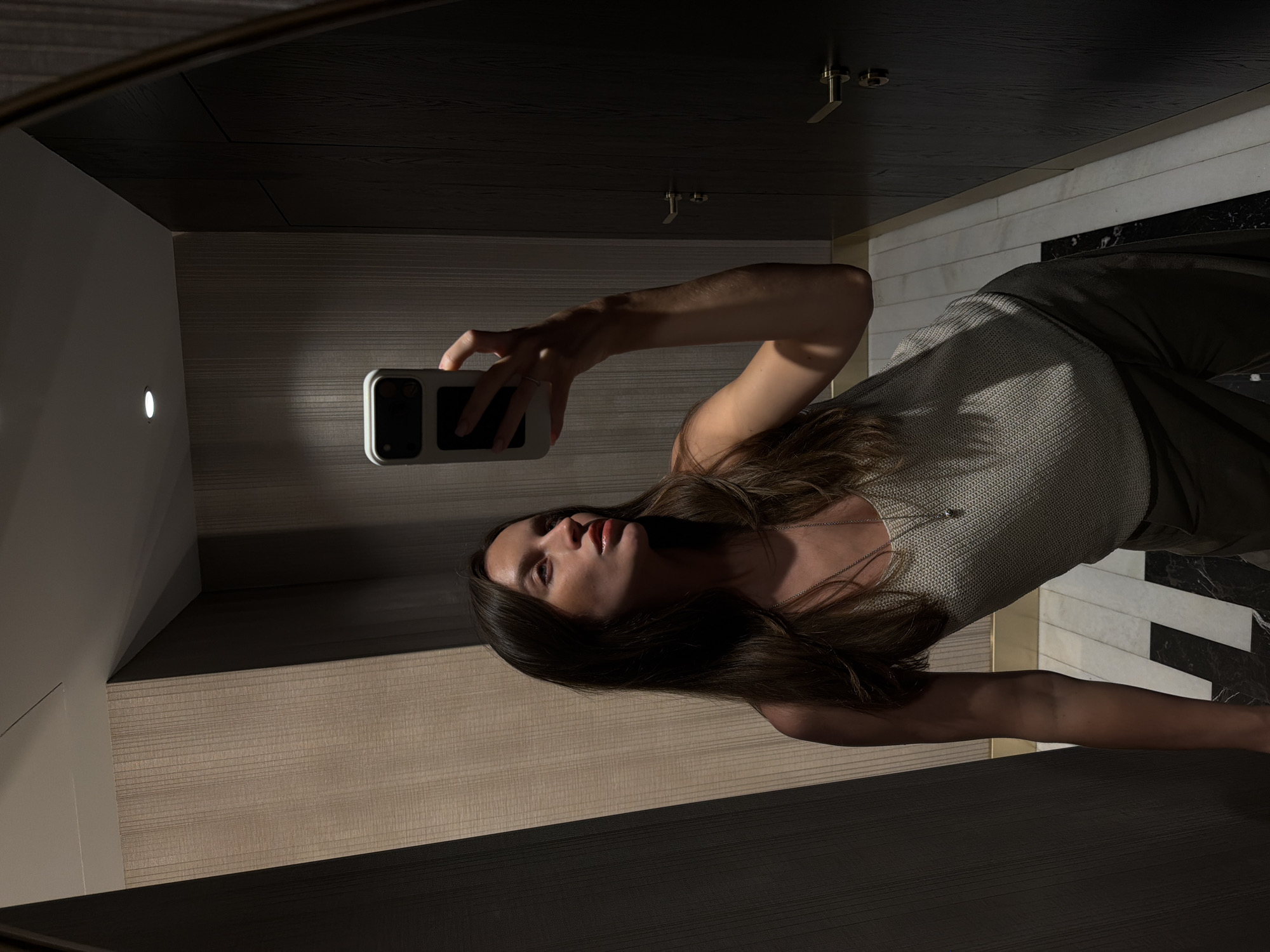 A woman is captured taking a mirror selfie in a dimly lit, modern interior space. She holds a white-cased smartphone in her right hand, extending her arm to frame the shot. Her head is tilted slightly, and she looks towards the phone, her lips appearing glossy. She has long, dark hair and wears a light-colored, possibly knit, top with a delicate necklace.
The reflection reveals a contemporary room with distinct architectural elements. The upper portion of the reflected background features dark, horizontal wooden paneling, while the lower section has light-colored, textured horizontal panels. A small, bright circular light source is visible on the light-textured wall. Brass-toned fixtures, possibly door handles or decorative elements, are mounted on the dark paneling. The floor visible in the reflection features a striking black and white geometric tile pattern. The lighting is characterized by strong contrasts, creating noticeable shadows on the woman's face and body, and emphasizing the textures of the room. There is no visible text in the image.
