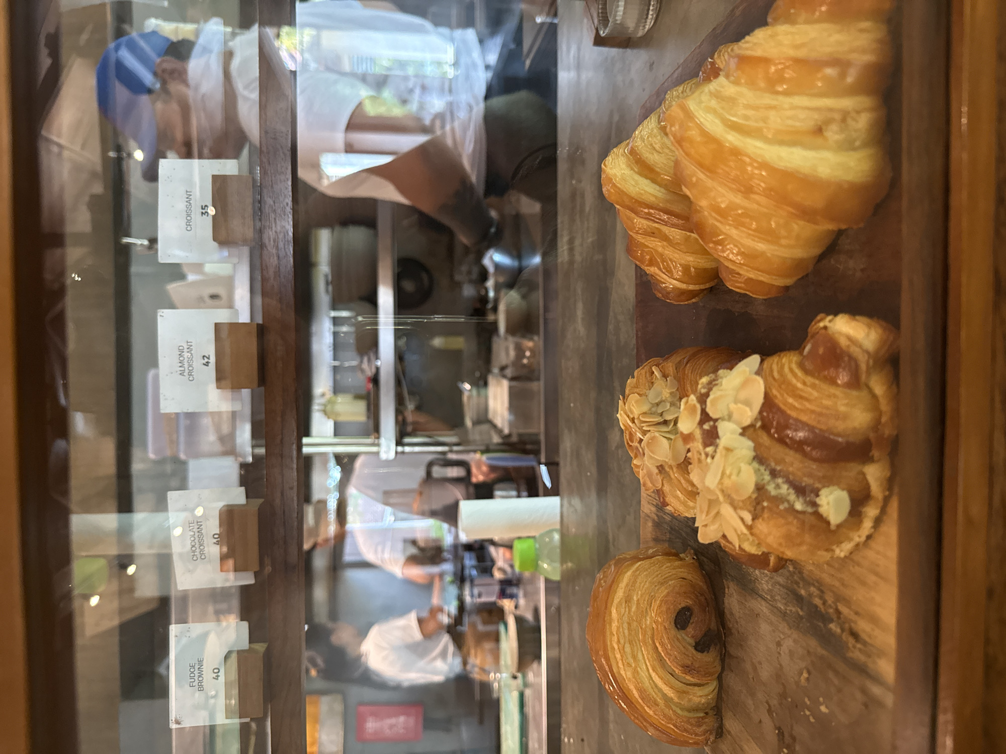 The image captures a close-up view of a pastry display case in what appears to be a bakery or cafe in Kuta Selatan, Indonesia. The scene is presented through clear glass, showing a selection of freshly baked goods arranged on a rustic wooden board.
In the foreground, on the right side of the frame, several pastries are visible. Two golden-brown, classic croissants with flaky layers are positioned at the top. Below them, an almond croissant is showcased, generously topped with sliced almonds and a dusting of what appears to be powdered sugar. At the bottom right, a round, swirled pastry, possibly a pain aux raisins or similar, with visible chocolate chips, completes the arrangement.
On the left side, behind the glass, are several white price tags on small wooden blocks, indicating the items and their prices. Visible text includes:
* "CROISSANT 35"
* "ALMOND CROISSANT 42"
* "CHOCOLATE CROISSANT 40"
* "FUDGE BROWNIE 40"
The glass also reflects the background, revealing the interior of the establishment. Reflections show at least two individuals, likely staff members, engaged in work. One person wears a blue cap, and another appears to be in a white uniform. Behind them, kitchen or preparation areas are dimly reflected, suggesting an active working environment. The overall lighting is bright, indicating it is likely daytime. The image focuses on showcasing the pastries while subtly hinting at the operational aspect of the cafe through the reflections.