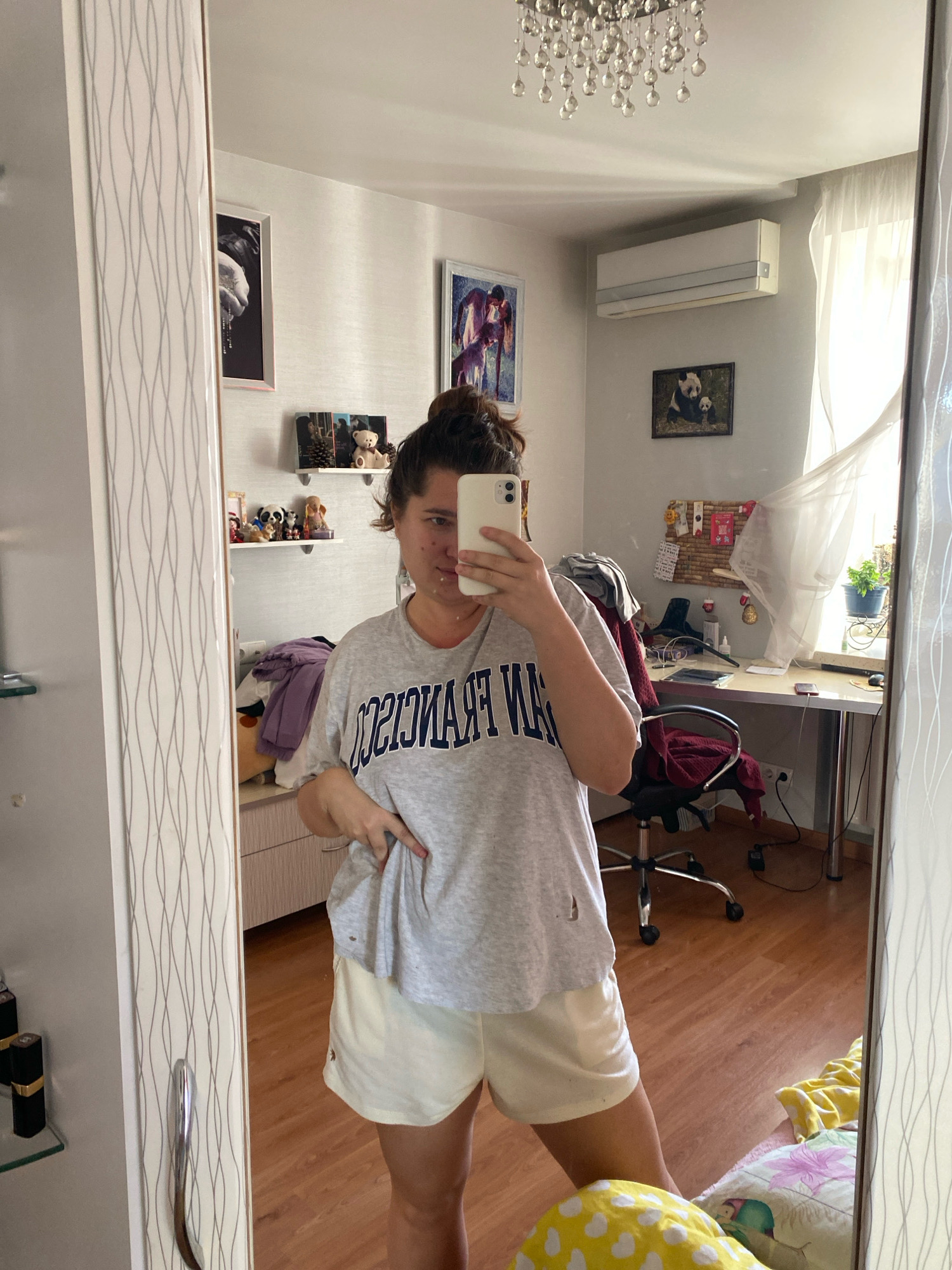 A young woman with her hair up in a bun is taking a mirror selfie in a room, likely a bedroom, as indicated by the bed with pillows and blankets visible in the reflection. She is wearing a grey t-shirt with "SAN FRANCISCO" written across it in navy blue letters and light yellow shorts. Her left hand is tucked into her waistband. The room is decorated with several framed pictures on the wall, including one of a panda and its cub and another that appears to be abstract art. There is a desk with a computer and office chair, and a window with sheer curtains. An air conditioner unit is mounted high on the wall. In the foreground, part of a mirrored wardrobe is visible, with shelves holding decorative items and some makeup. The flooring is wooden.