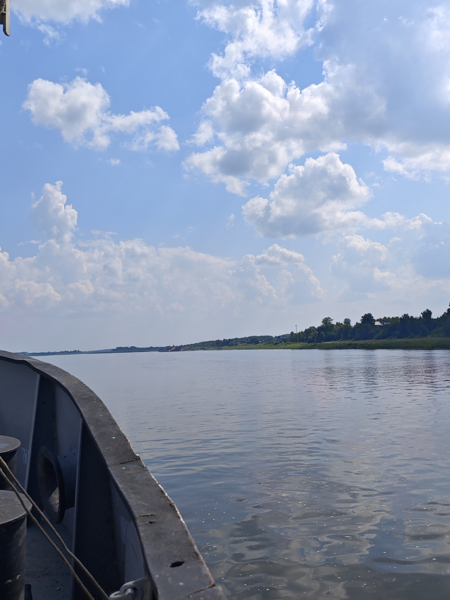 A wide, calm river stretches towards a distant, tree-lined horizon under a bright blue sky filled with fluffy white clouds. The foreground features the dark, weathered edge of a vessel, with mooring posts visible. The riverbank on the right side of the image is densely wooded, with glimpses of scattered houses and a few taller structures amongst the trees. The water reflects the sky and the distant landscape, with subtle ripples indicating a gentle current. The scene evokes a sense of peaceful travel on a sunny day.