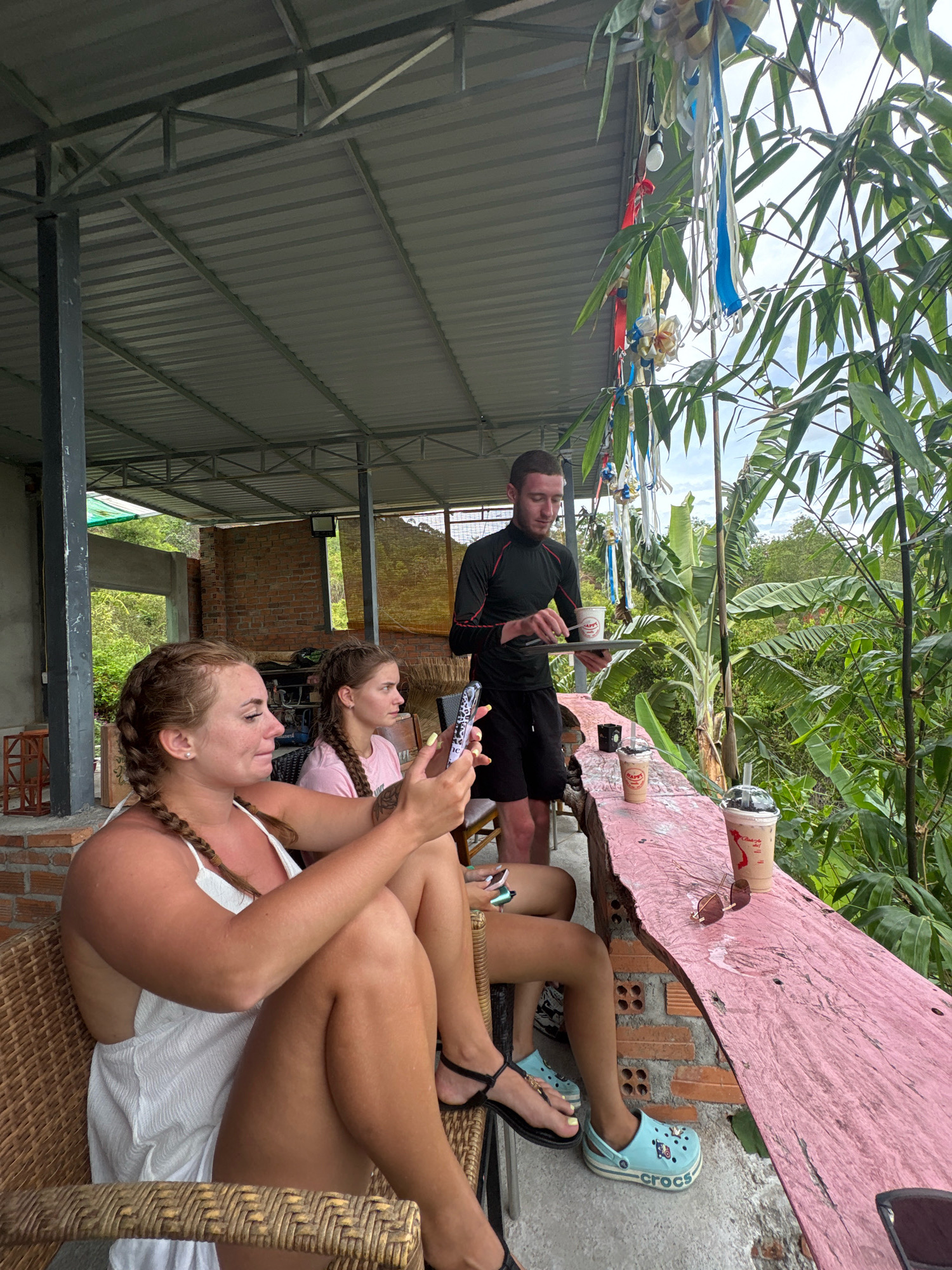 In a cafe in Nha Trang, Vietnam, three people are present. The foreground features two women sitting at a table, one engrossed in her phone, while the other looks off into the distance. Behind them, a man in a black long-sleeved shirt and shorts serves drinks. The cafe has an open-air seating area with a corrugated metal roof and a wooden railing overlooking a lush green landscape. The table is made of rough-hewn pink wood, and several drinks are placed on it. The overall atmosphere suggests a relaxed setting, possibly a tourist spot given the tropical foliage and open-air design.