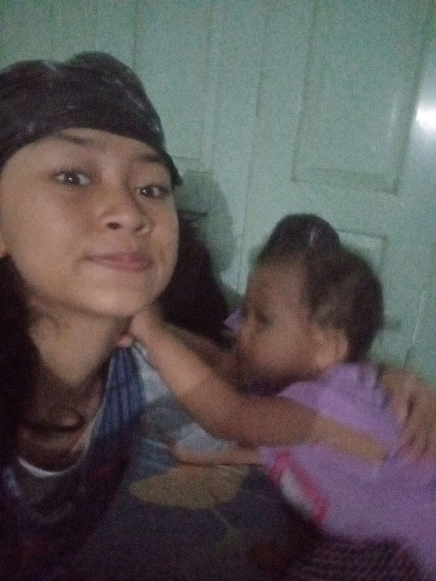 This is a close-up, slightly blurry selfie taken by a young woman wearing a dark bandana. She is looking directly at the camera with her hand touching her chin. To her right, a baby wearing a purple shirt is visible, with its arm reaching towards the woman's neck. The background appears to be a light green wall with some paneling. The overall lighting is dim, suggesting the photo was taken indoors. The scene is likely a home environment.