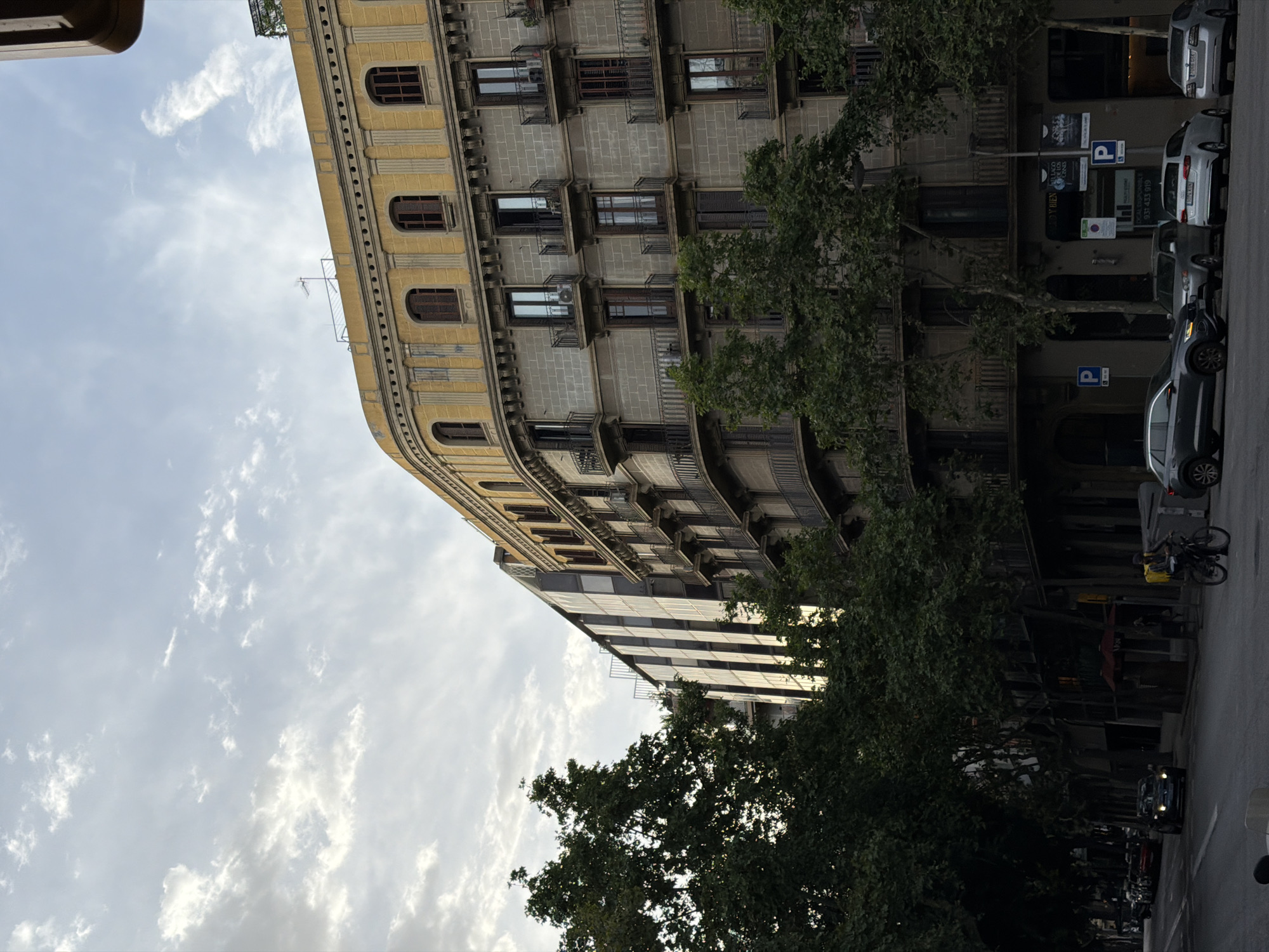 The image captures a street scene in Barcelona, Spain, featuring a grand, old building with a curved facade and a yellow top floor adorned with arched windows. This building is juxtaposed with a modern, glass-fronted structure to its left. Lush green trees line the street, partially obscuring the lower levels of the buildings and casting shadows on the road. The street is paved, and several cars are visible, some parked and others in motion. A person on a bicycle is also present, riding on the sidewalk. The sky above is partly cloudy, with soft light filtering through. The architecture and tree-lined street suggest a vibrant urban environment.