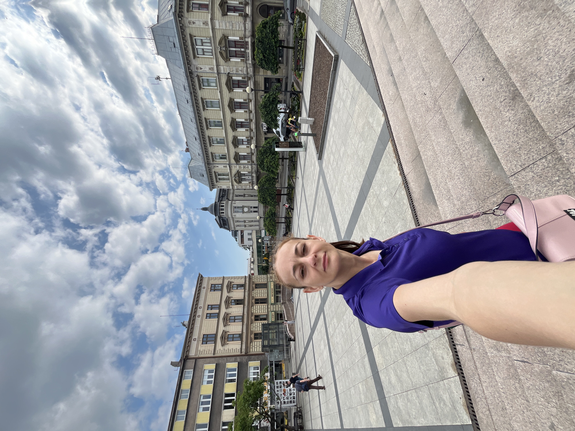 A woman in a purple shirt takes a selfie in a city square. She is standing on a wide, stone staircase, with the square stretching out before her. The square is paved with grey tiles and has several buildings surrounding it. One building has a yellow facade, while others are adorned with classical architectural details. Trees are planted in rows along the perimeter of the square, and there are benches and streetlights. The sky is partly cloudy. In the background, people are walking and sitting in the square. There is also a white van and a car visible.