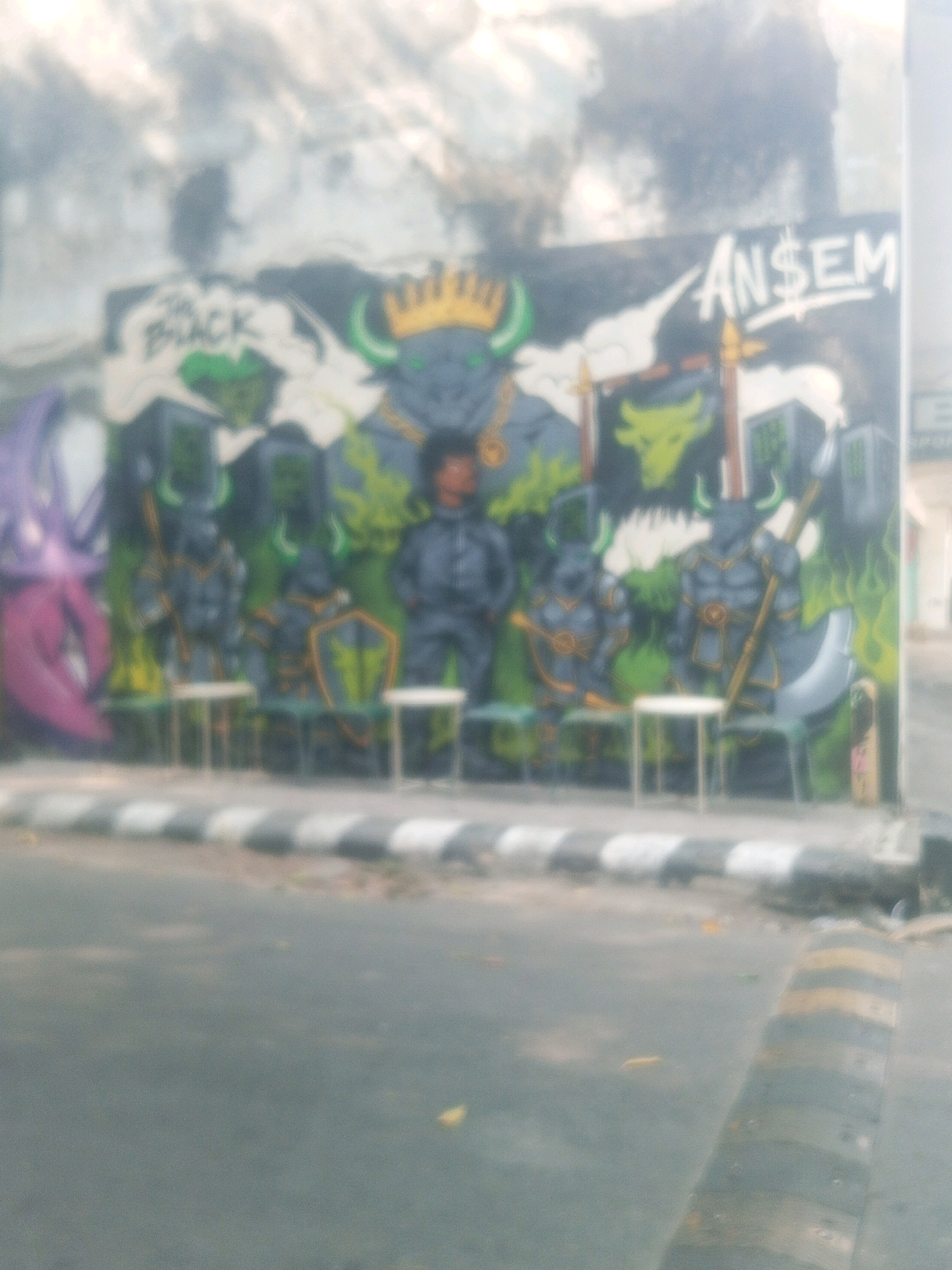 The image depicts a mural on a wall, likely in an urban setting. The mural features stylized bull figures wearing armor and crowns, suggesting a theme of strength or a mythical narrative. Some bulls have green horns and wear golden necklaces. One prominent bull figure has a gold crown. To the left of the bull figures, a purple, abstract, possibly mythological creature is visible. Text in white lettering is present on the mural; "THE BLACK" is seen on the left, and "AN$EM" with a dollar sign replacing the 'S' is on the right. In front of the mural, there are several small white stools or tables arranged, hinting at an outdoor seating area. The background is blurry, but appears to be a wall with some sky or clouds. The foreground shows a road with a pedestrian crossing or speed bump pattern, indicating a public thoroughfare. The overall scene is somewhat blurred and the lighting suggests it might be daytime. Based on the provided information that the location is Kediri City, Indonesia, the mural is likely a piece of street art found within the city.