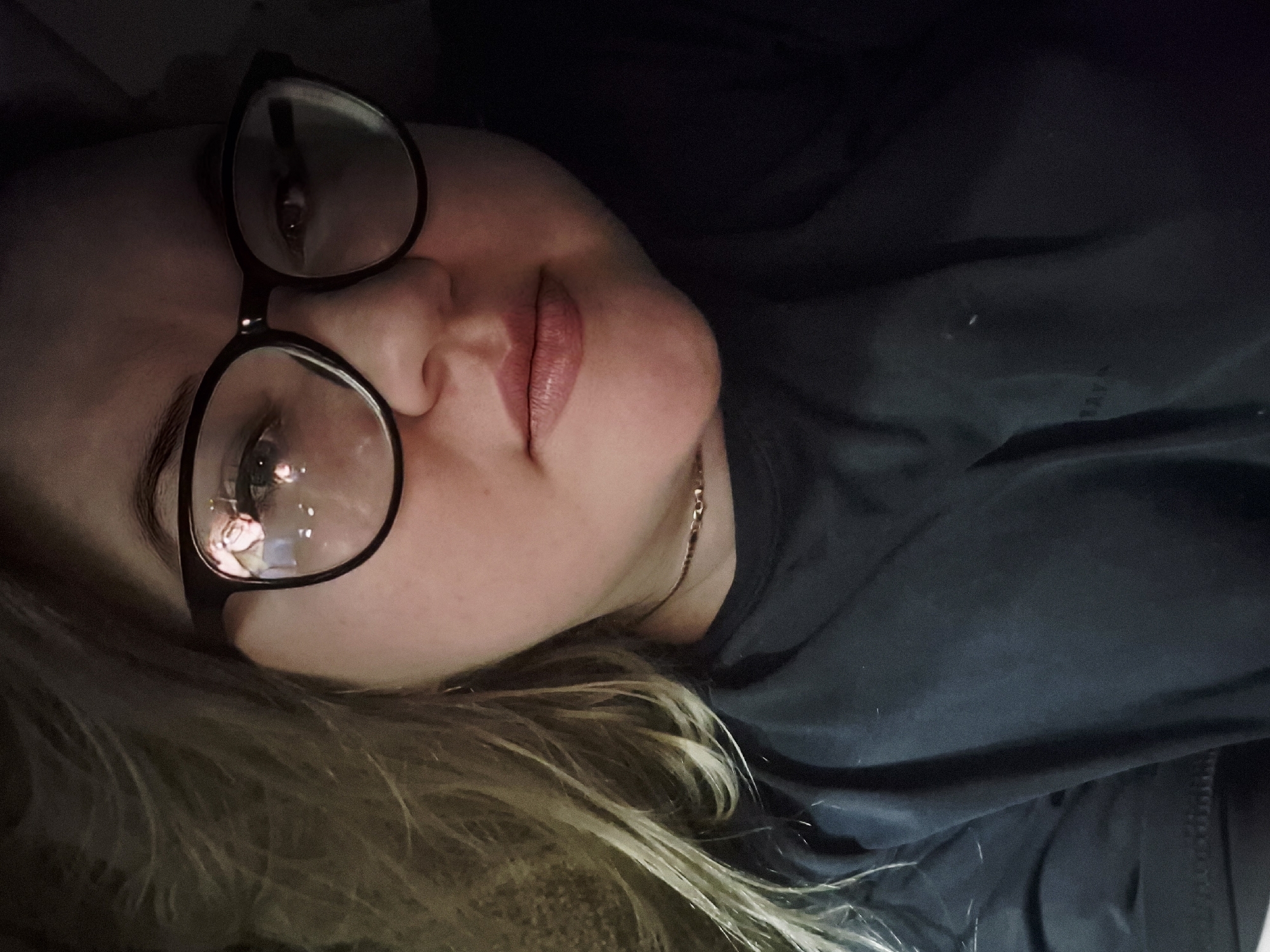 A close-up portrait, slightly angled, depicts an adult female positioned within what is identified as the city of Фоки, Russia. The subject has fair skin and a round facial structure, with light-colored, wavy hair visible on the left side of the frame. She wears dark-framed, circular eyeglasses, which display reflections of multiple small, bright light sources from an unidentifiable internal environment. Her gaze is directed straight forward. A thin, metallic choker-style necklace adorns her neck, and she is dressed in a plain, dark-colored, short-sleeved garment. Her lips are naturally colored and slightly parted. The background is uniformly dark and out of focus, suggesting a dimly lit indoor setting. The overall lighting is subdued, appearing to originate from in front of the subject.