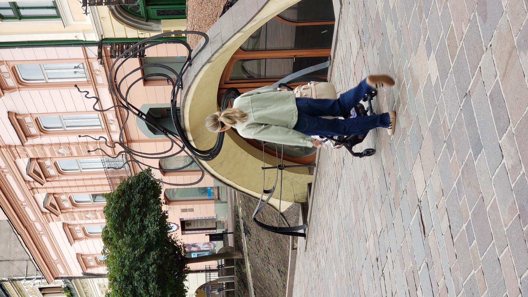 A rear view medium shot captures a person walking a stroller across a paved plaza in Prešov, Slovakia. The person is wearing a light green jacket and dark blue jeans, with blonde hair pulled back in a ponytail. They are pushing a black stroller with a beige bag attached to the handle. In the background, a light pink building with ornate windows and architectural details stands. In front of the building is a unique, curved structure with black, winding metalwork resembling antennae. A green tree with dense foliage is to the left of the building. The plaza is paved with rectangular, multi-colored stones. The scene appears to be in an urban environment, likely a pedestrian area.