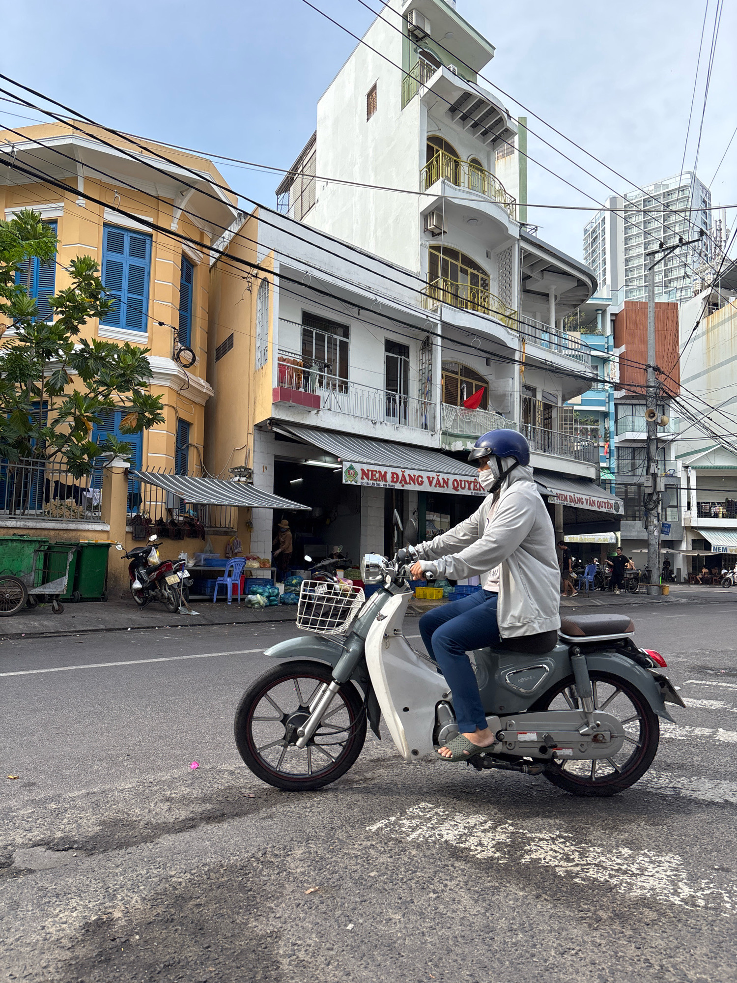A person wearing a blue helmet, face mask, light gray jacket, and dark blue pants rides a grey scooter on a paved city street in Nha Trang, Vietnam. The scooter has a white front and a basket. In the background, there are buildings with yellow and white facades, some with blue shutters, and overhead wires. A few motorcycles and trash bins are visible near a storefront with a striped awning. The street is wet in patches, and there are shadows cast on the pavement. The sky is partly cloudy.