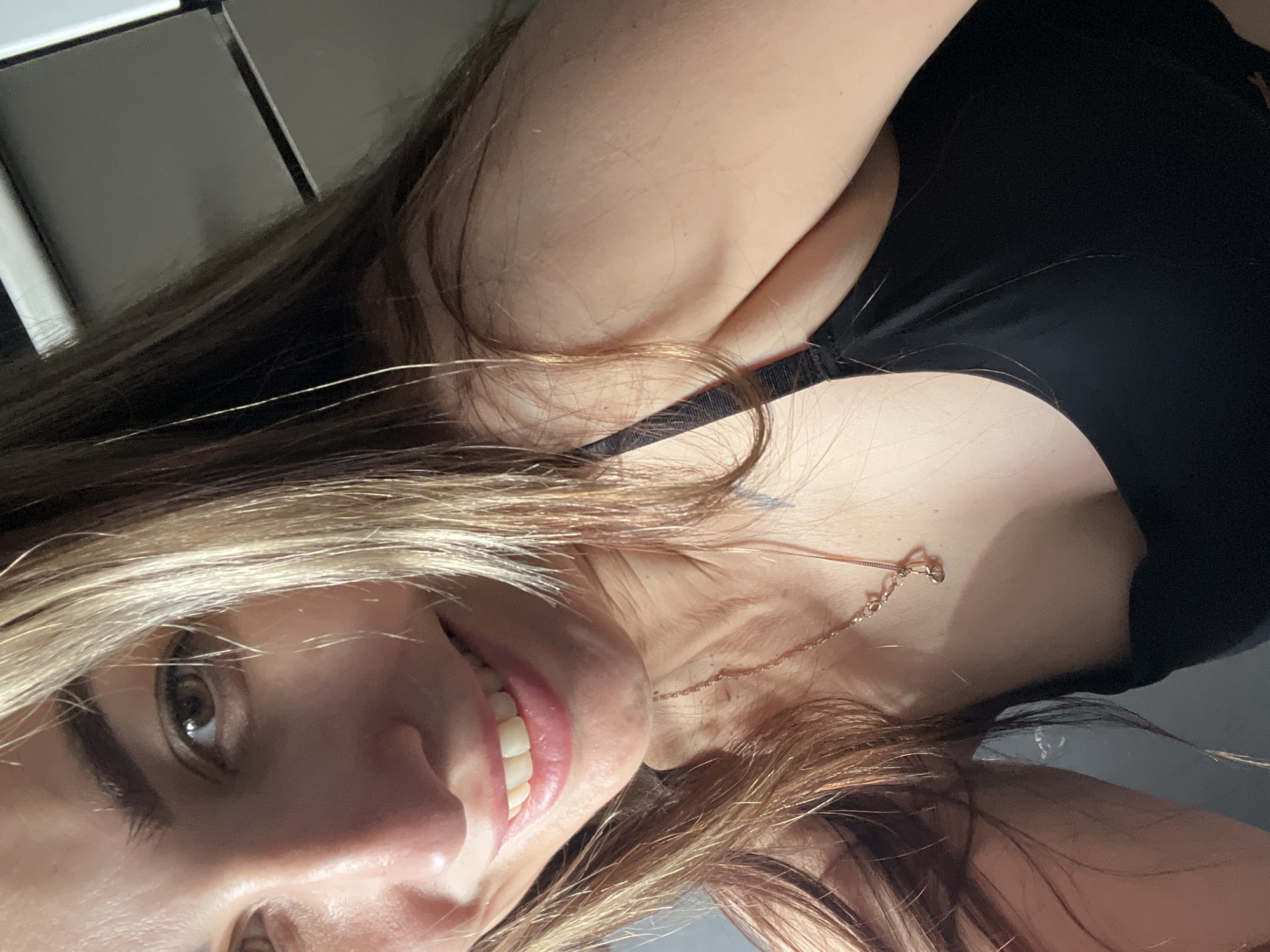 A close-up, tilted image features a person with fair skin, light brown hair with blonde highlights, and a black top. A delicate, gold-colored necklace rests on the person's neck. The background is blurred, showing hints of gray and white surfaces. The person is smiling, revealing white teeth. The setting is unspecified.