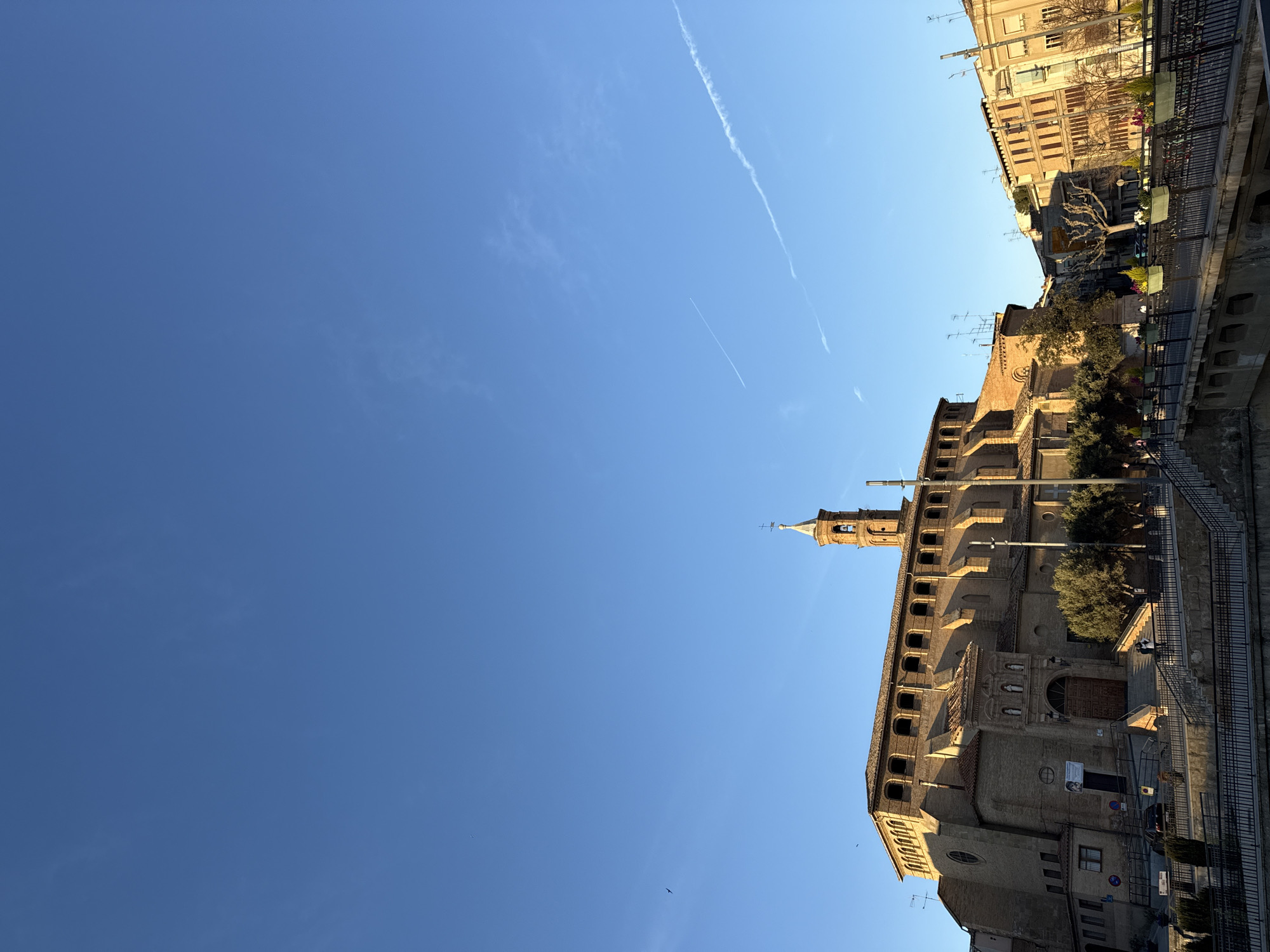 This image presents a detailed view of a city streetscape in Barbastro, Spain, under a clear, bright blue sky. The primary subject is a large, multi-story brick building, identifiable as a cathedral or basilica, featuring a prominent, narrow bell tower topped with a pointed spire and a cross. The building's facade is characterized by a series of arched windows along an upper loggia and a grand main entrance adorned with statuary, including three figures positioned above the archway. The structure is illuminated by warm, low-angle sunlight, casting defined shadows.
In the foreground, a bridge with dark metal railings and decorative planter boxes containing various plants extends across the lower portion of the frame. Small human figures are discernible on the bridge and ascending a set of stairs leading to the cathedral's entrance. A single vehicle is parked near the base of the cathedral. To the right of the central edifice, other multi-story urban buildings with windows and balconies are visible, also bathed in direct sunlight. Scattered trees, some with bare branches and others evergreen, are present within the scene. The upper two-thirds of the image consist of a vibrant blue sky, featuring several faint, linear contrails from aircraft and a single small bird in flight.