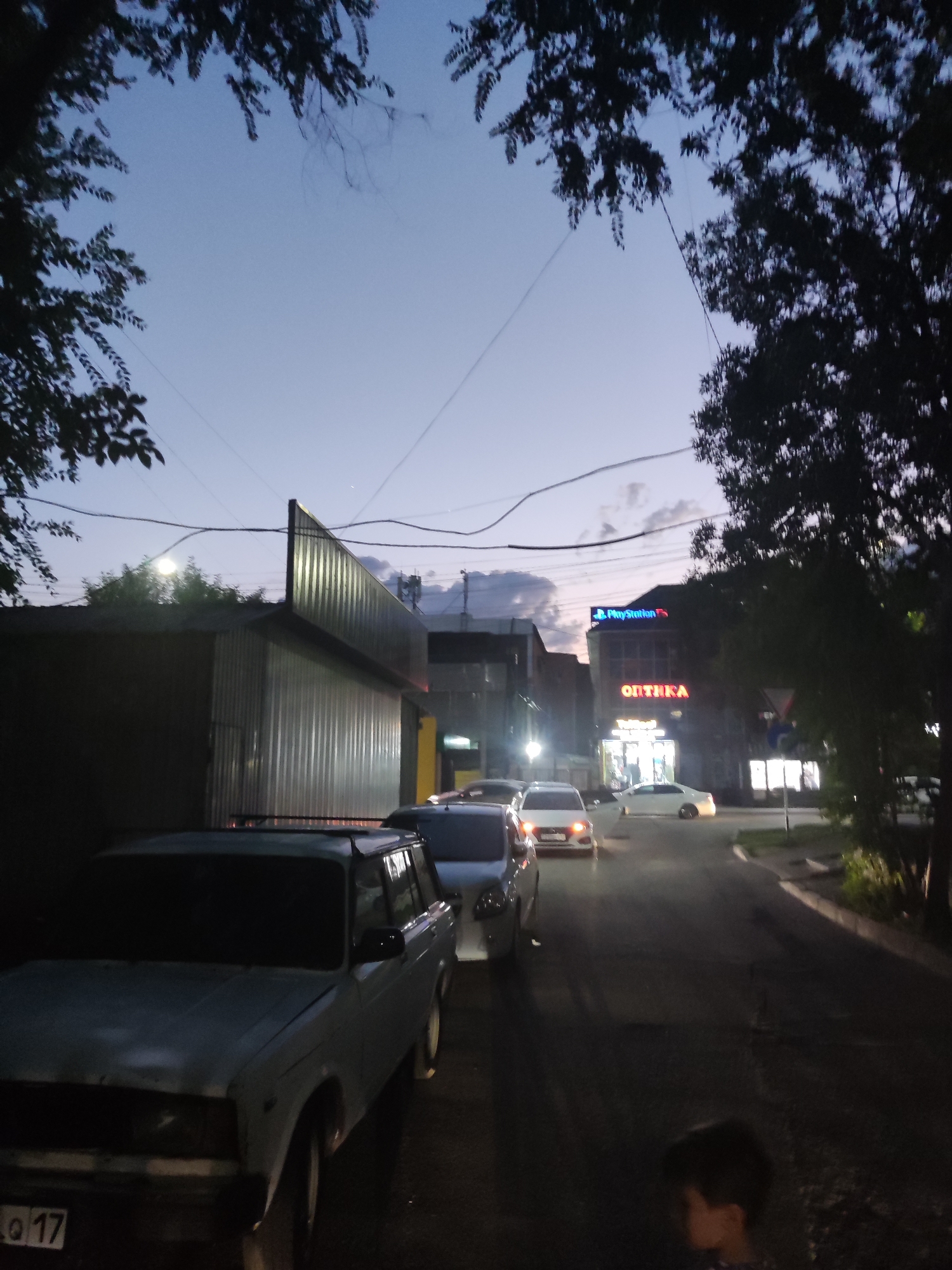 The image depicts a street scene at dusk in Shymkent, Kazakhstan. A light blue, older model car is parked on the left side of the street, with its license plate visible showing "0 17". Further down the street, other cars are also parked or driving. On the right side of the street, a building features illuminated signs, including one that reads "PlayStation" in blue neon lights and another with red lettering that appears to be "ОПТИКА" (Optika, meaning optics). The sky is a pale blue, transitioning to darker hues as night approaches, with some clouds visible. Trees with dark foliage frame the top of the image, and electrical wires crisscross the sky. In the foreground, a portion of a person's head and shoulders is visible, suggesting they are walking or standing in the street. The overall atmosphere is that of a typical urban street scene in the early evening.
