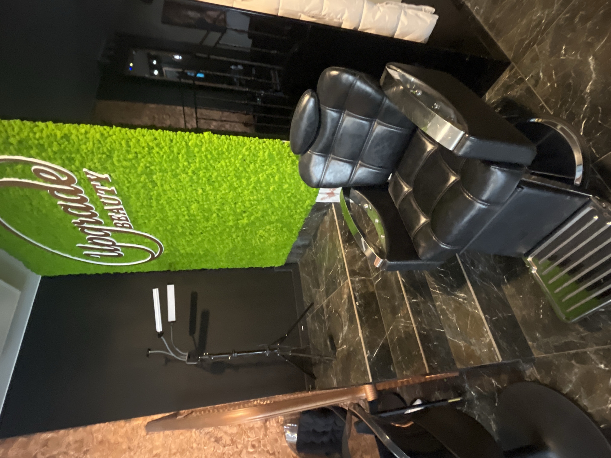 The image depicts the interior of a beauty salon. A black leather reclining chair with a chrome footrest is positioned in the foreground. Behind the chair, a wall is covered with a vibrant green moss installation, featuring the words "Upgrade Beauty" in white lettering. To the left of the moss wall, a black tripod with two LED light bars is visible, casting shadows on the black wall. A tall, ornate mirror with a copper-toned frame stands next to the tripod. The floor is made of dark marble tiles with white veining. The background shows a staircase leading up to a darker area, with what appears to be more salon seating and a white quilted wall covering. The lighting is dim, with the exception of the LED lights and the bright moss wall. The scene is clean and modern, suggesting a professional service environment. The location is stated to be Białystok, Poland. There are no people visible in the image.