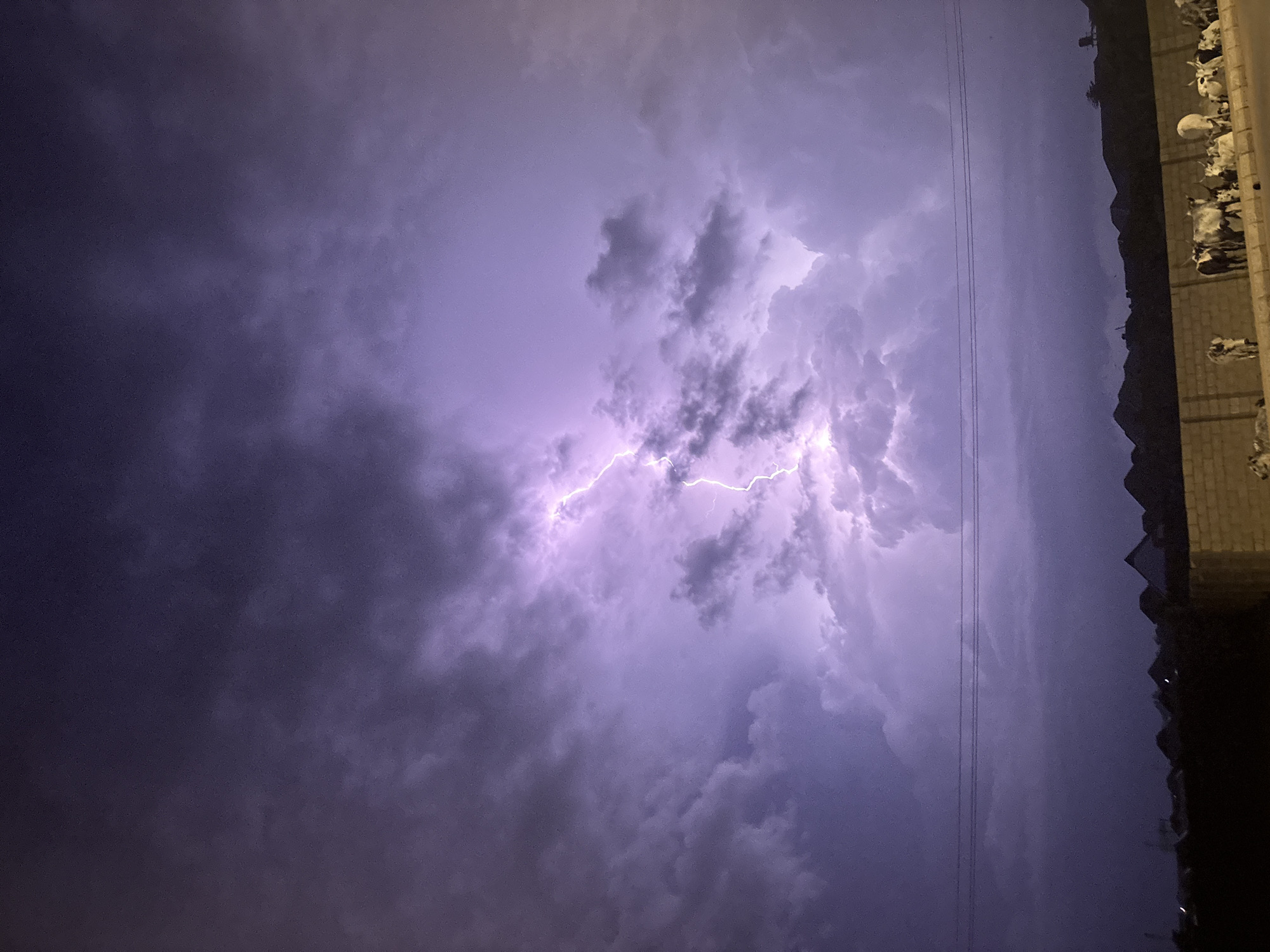 The image captures a dramatic nighttime scene in Bukuru, Nigeria, dominated by a powerful lightning strike illuminating the dark, turbulent sky. The lightning, a jagged bolt of bright white light tinged with purple, cuts through dense, swirling clouds. These clouds exhibit varying shades of purple and grey, with some areas appearing almost black and others illuminated by the electrical discharge.
In the foreground, a low brick wall runs horizontally across the frame, with a road or pathway behind it. A herd of cattle are gathered on the other side of the wall, some standing and others lying down. The cattle appear to be of various breeds and colors, mostly white and brown. A few human figures, dressed in dark clothing, are visible standing amongst the cattle, seemingly observing the storm. Beyond the wall and the cattle, a silhouetted landscape of hills and what appear to be rooftops of buildings can be seen against the dimly lit sky. Power lines are visible as thin horizontal lines spanning the mid-ground of the image. The overall impression is one of a powerful natural event occurring in a residential or rural outskirts setting.