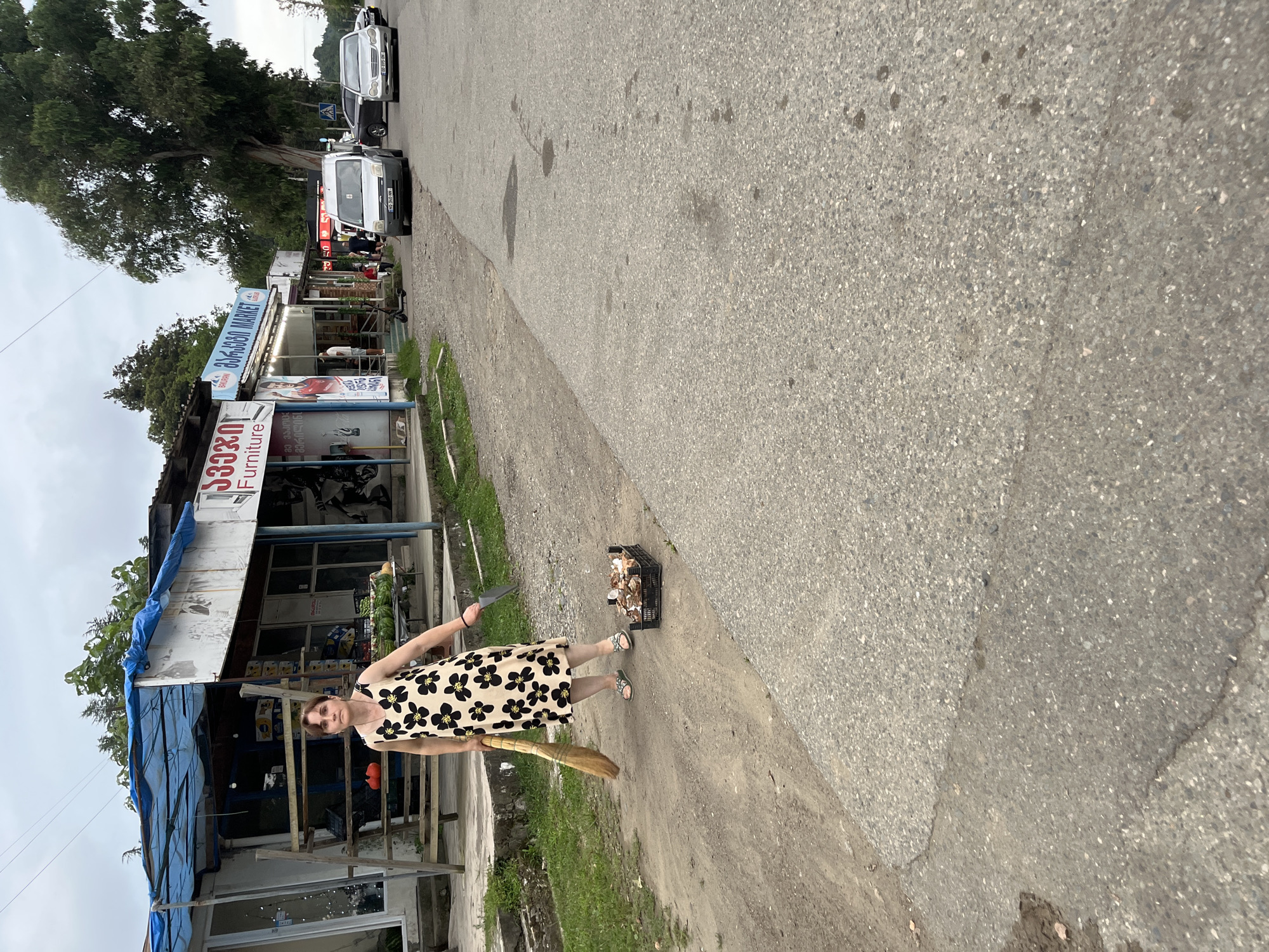 A woman in a floral dress is standing on a dirt and asphalt road in front of a row of small shops. She is holding a broom in her right hand and a knife in her left. To her right, there is a black crate filled with mushrooms. In the background, there are several vehicles and more shops, some with awnings and signs in Georgian. The sky is overcast. The scene appears to be in a semi-urban or rural setting.