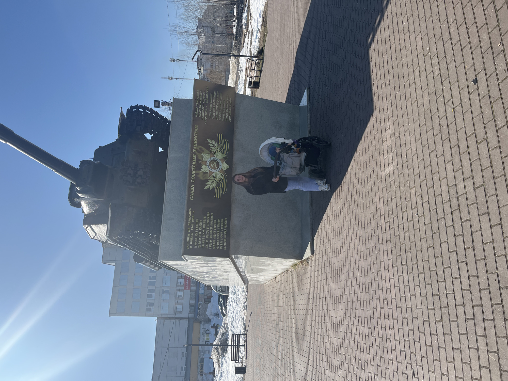 The image shows a memorial in Chaikovsky, Russia, featuring a Soviet-era tank mounted on a concrete base. A woman and a child in a stroller are standing in front of the monument.
The inscription on the concrete base reads "СЛАВА СОВЕТСКИМ ТАНКИСТАМ" (Glory to Soviet Tankers). A plaque lists names of war heroes. In the background, there are buildings, trees without leaves, patches of snow, benches, and power lines. The ground is paved with bricks. The sky is clear.