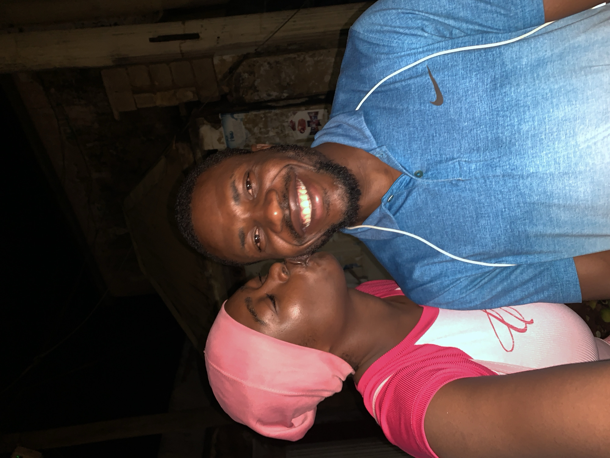 Two people are cheek to cheek and close to kissing. The person on top is a Black male wearing a light blue collared shirt with a Nike swoosh. He has short, dark hair and a beard. The person on the bottom has dark skin and is wearing a pink head covering and a pink and white shirt. The location appears to be a poorly lit indoor or covered outdoor area with brick walls or structures and possible overhead beams. The photo was taken in Kabong, Nigeria.