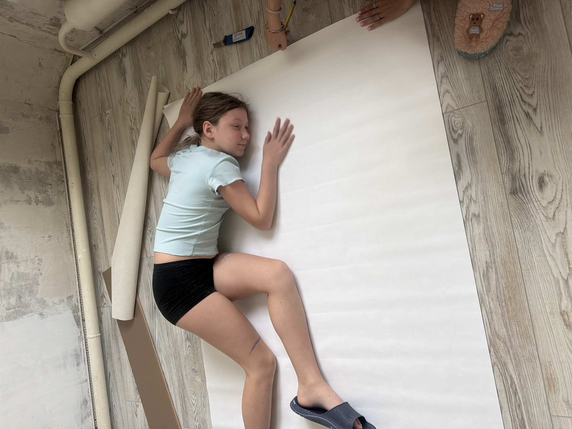 A young girl lies on a large sheet of white paper on a wooden floor. She wears a light blue short-sleeved shirt and black shorts. Her eyes are closed and she is lying on her side with both arms extended and hands touching the paper. A blue slipper is on her right foot. A darker blue mark is visible on her right leg.
A woman's hands are visible at the top of the paper, one holding a pencil. A utility knife is also placed on the paper.
Nearby are rolls of material. A pipe runs along the top left corner of the room. The wall near the pipe is unfinished.
No specific details in the image indicate a location in Markovo, Russia.