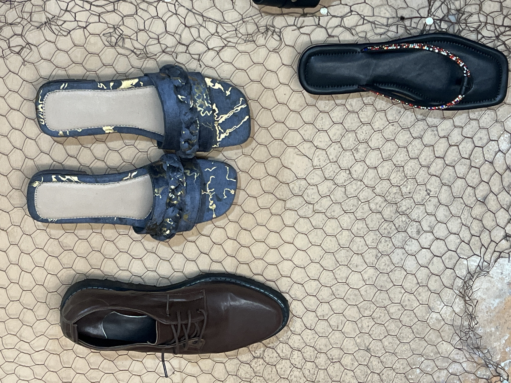 The image depicts a selection of footwear arranged on a surface covered with a hexagonal wire mesh. There are two pairs of blue slides with gold accents, a black bejeweled flip-flop, and a pair of brown leather lace-up shoes. The background is a light beige or gray, possibly a wall or floor. There are no people visible. The scene is possibly located in Jos, Nigeria.