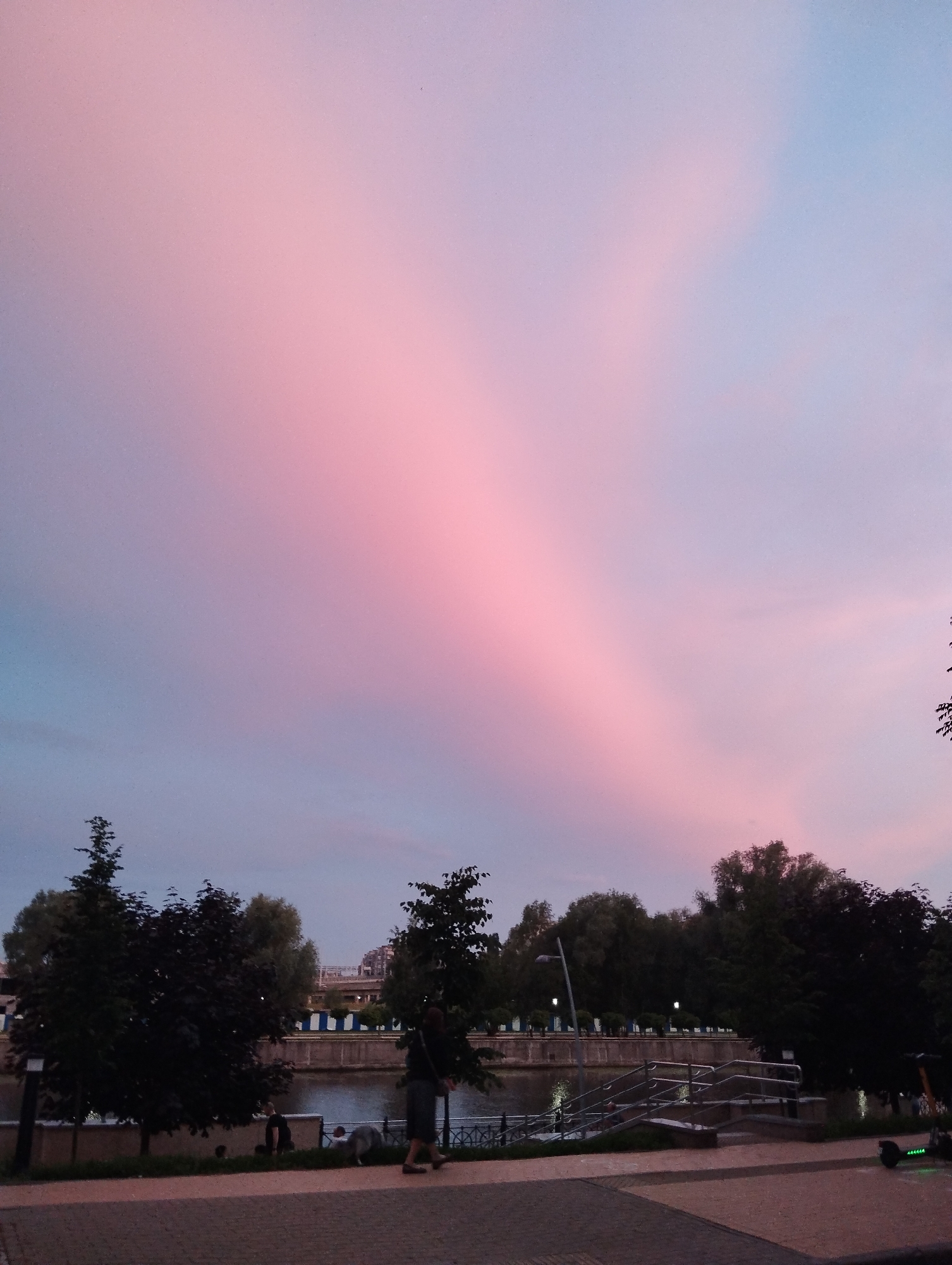 The image depicts a tranquil evening scene in Kaliningrad, Russia. The sky is a prominent feature, awash with soft hues of pink and blue, suggesting either sunrise or sunset. A band of pink light stretches diagonally across the sky, creating a dramatic and ethereal effect.
The foreground shows a paved walkway with a cobblestone pattern. To the left, silhouetted trees are visible, their dark forms contrasting with the lighter sky. In the middle ground, a river or canal separates the walkway from the opposite bank. Along the far bank, a line of trees forms a backdrop, with some buildings visible behind them. A metal railing with steps leads down towards the water.
Several people are present in the scene, though their features are not clearly discernible due to the low light. One person is walking along the pathway, and another appears to be near the water's edge. A dog is also visible walking alongside a person. An electric scooter is parked on the right side of the walkway, its green lights illuminated.
The overall mood is peaceful and serene, enhanced by the soft lighting and the natural beauty of the sky. The time of day is clearly evening or early morning, as indicated by the sky colors and the artificial lights that are beginning to switch on. The presence of a river and trees suggests an urban park or waterfront area. There is no visible text in the image.