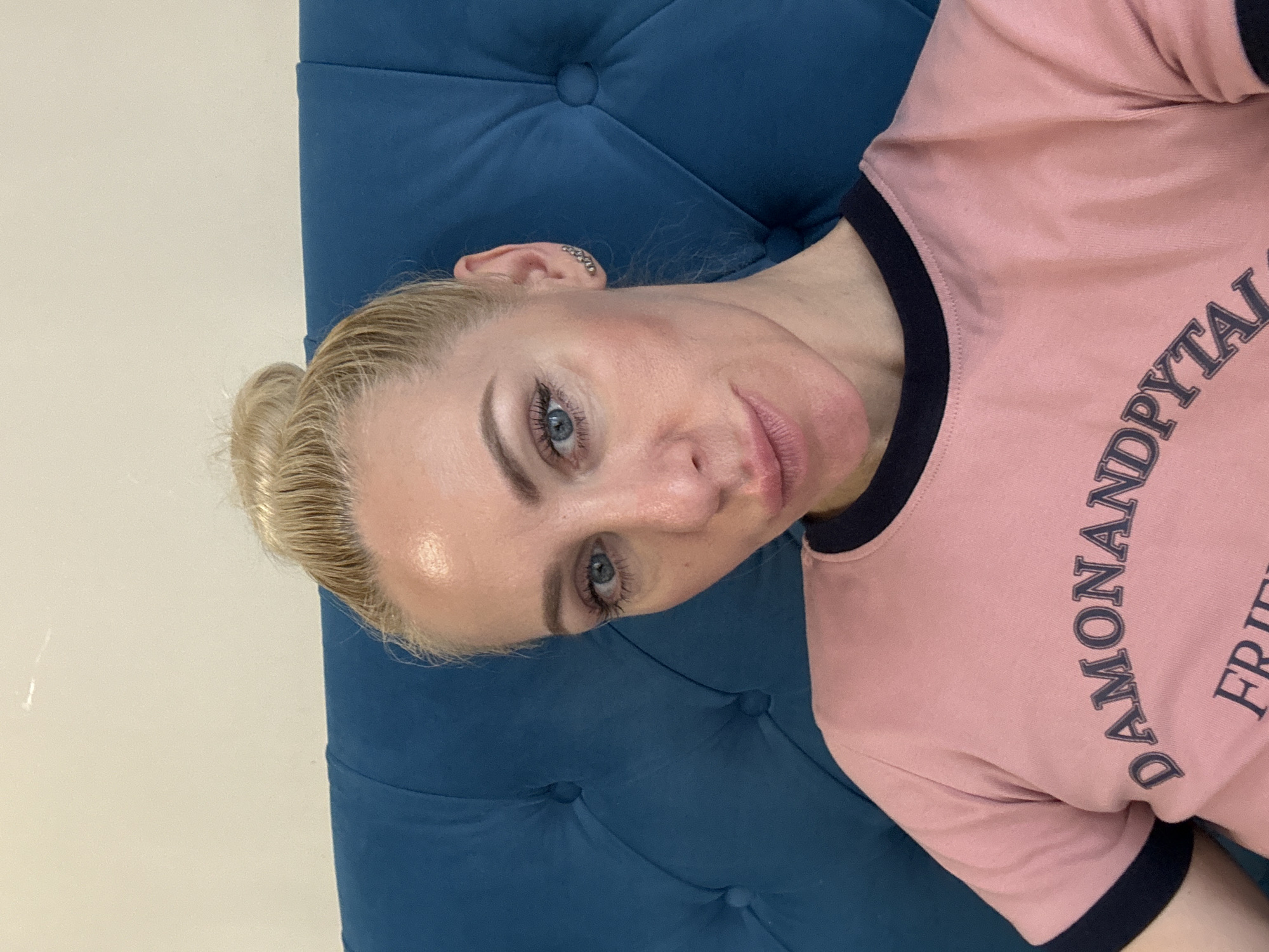 A blonde woman with her hair in a bun is pictured from the chest up. She is wearing a pink t-shirt with a dark blue trim around the collar and sleeves. The t-shirt has text on it, but only the letters "DAMONAN DP" and "FR" are fully visible. She is wearing a small silver earring on her right ear. The woman is leaning against a tufted, dark blue headboard. The background is a plain, light-colored wall with some minor marks. The image appears to be a selfie taken indoors. The city of Unknown, Russia is not depicted or suggested in the image.